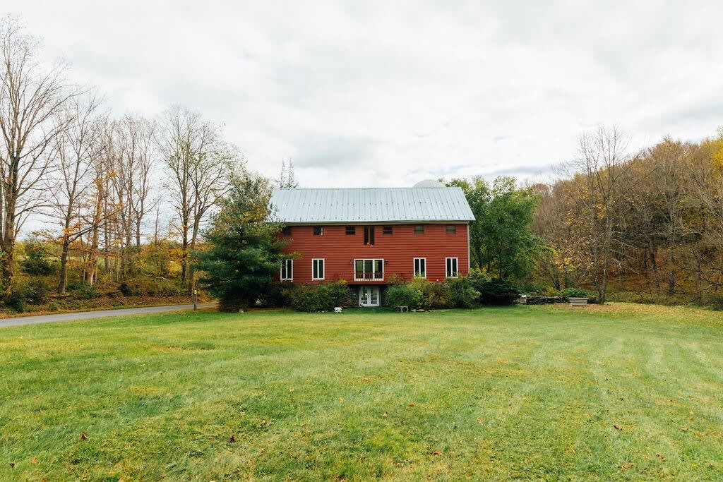 Front view of the red barn house with a metal roof, surrounded by a large green lawn and autumn trees. The house features multiple windows and a central entrance, with a road visible to the left.