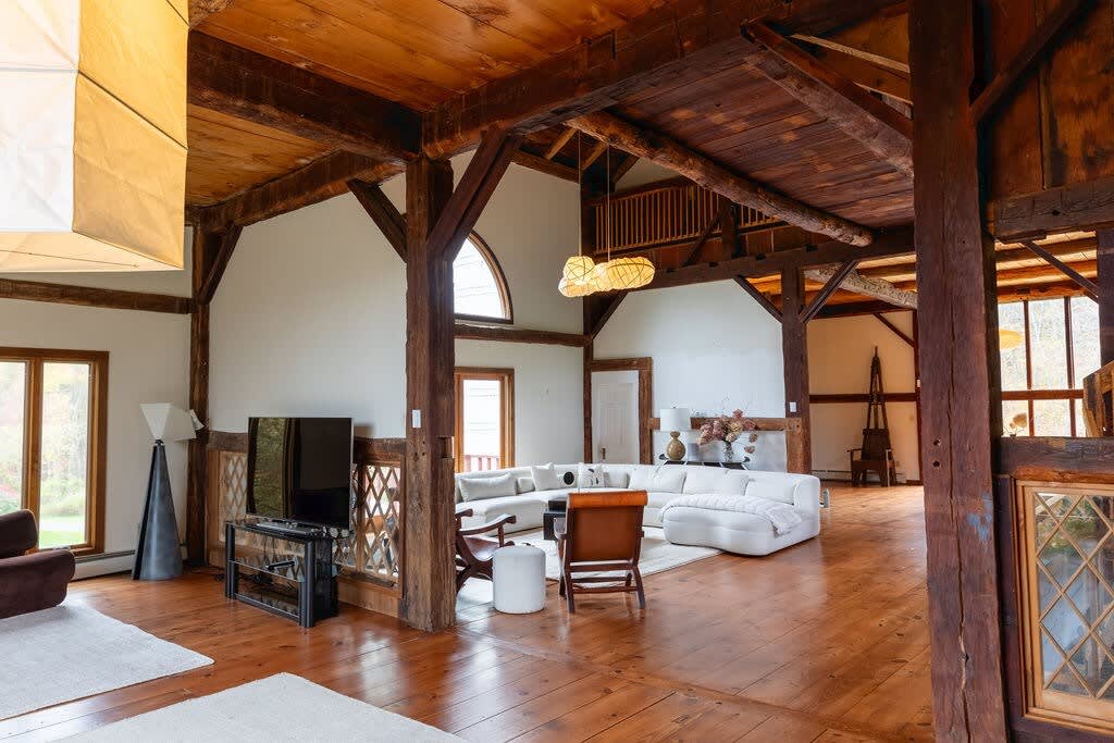 Expansive living area with warm hardwood floors and exposed wooden beams, featuring a white sectional sofa on a light rug and a separate curved white sofa. A large flat-screen TV is on a stand, and a unique floor lamp adds a modern touch to the rustic setting.
