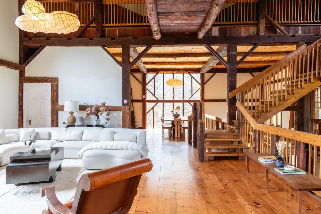 Open-concept living space with a white sectional sofa on a light rug, facing a dark coffee table, all set on warm hardwood floors. Exposed wooden beams and a staircase define the rustic barn aesthetic, with a dining area visible through large windows in the background.