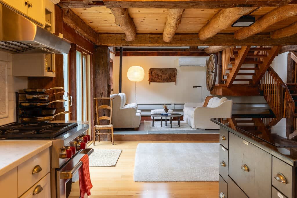 An open-concept living space featuring a modern kitchen island in the foreground, leading to a cozy seating area with white armchairs and a rustic wooden staircase, all under exposed wooden beams.