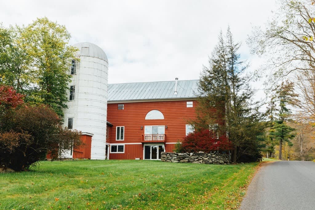 The striking red barn-style house with a tall white silo attached, surrounded by a lush green lawn and vibrant autumn trees, with a paved road leading up to the property.