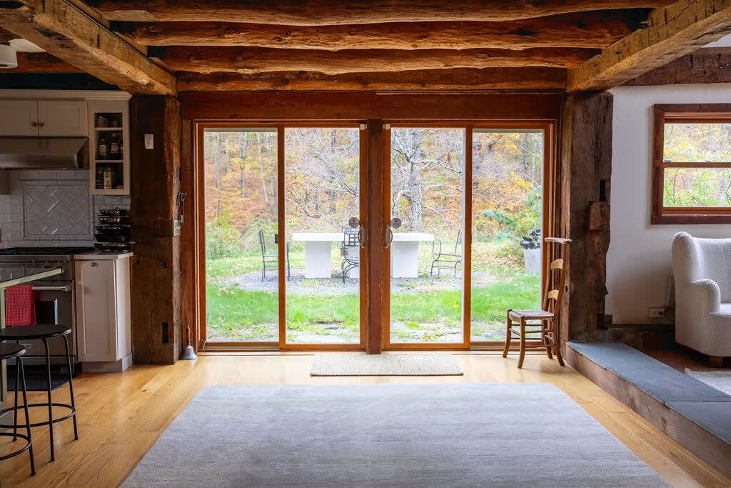 An open living space with light hardwood floors and prominent exposed wooden beams, featuring large glass sliding doors that offer a picturesque view of a grassy backyard with outdoor furniture.