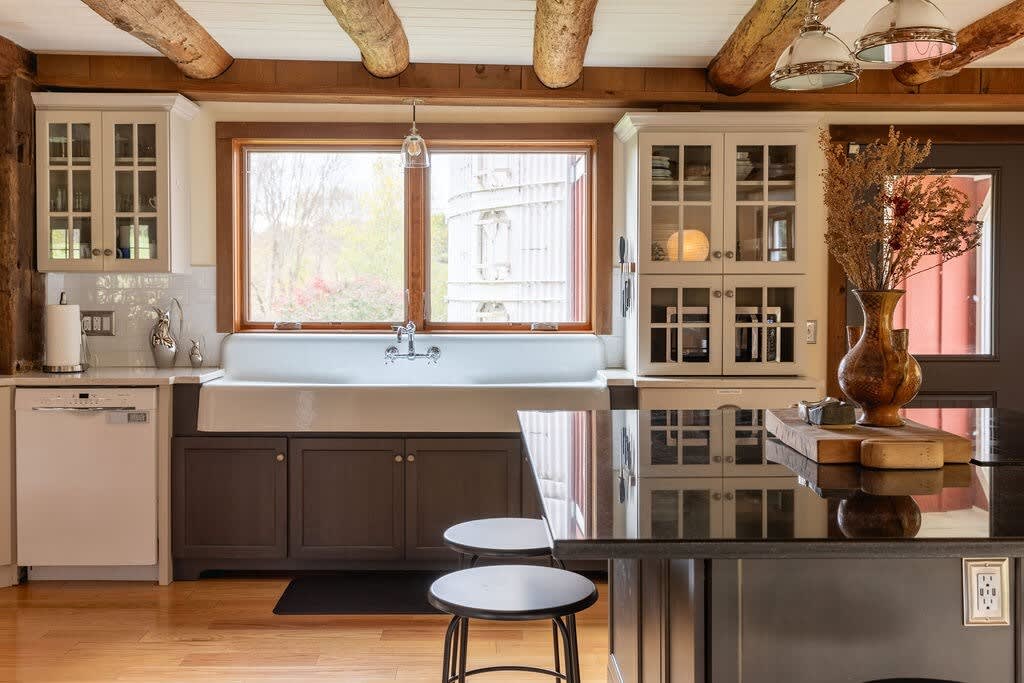 A rustic-modern kitchen featuring a large white farmhouse sink under a window, complemented by white upper cabinets, dark lower cabinets, and a black granite island with bar stools.
