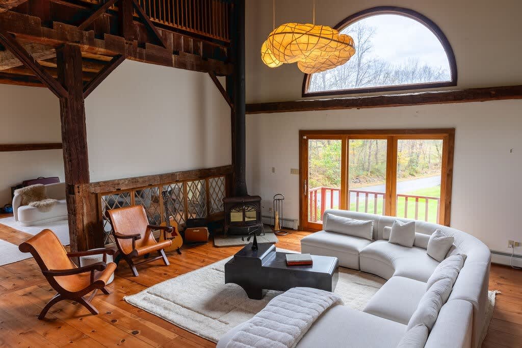 Cozy living room featuring a large white sectional sofa on a light rug, centered around a dark, sculptural coffee table. A black wood-burning stove stands in the corner, beneath an arched window, all framed by exposed wooden beams and warm hardwood floors.