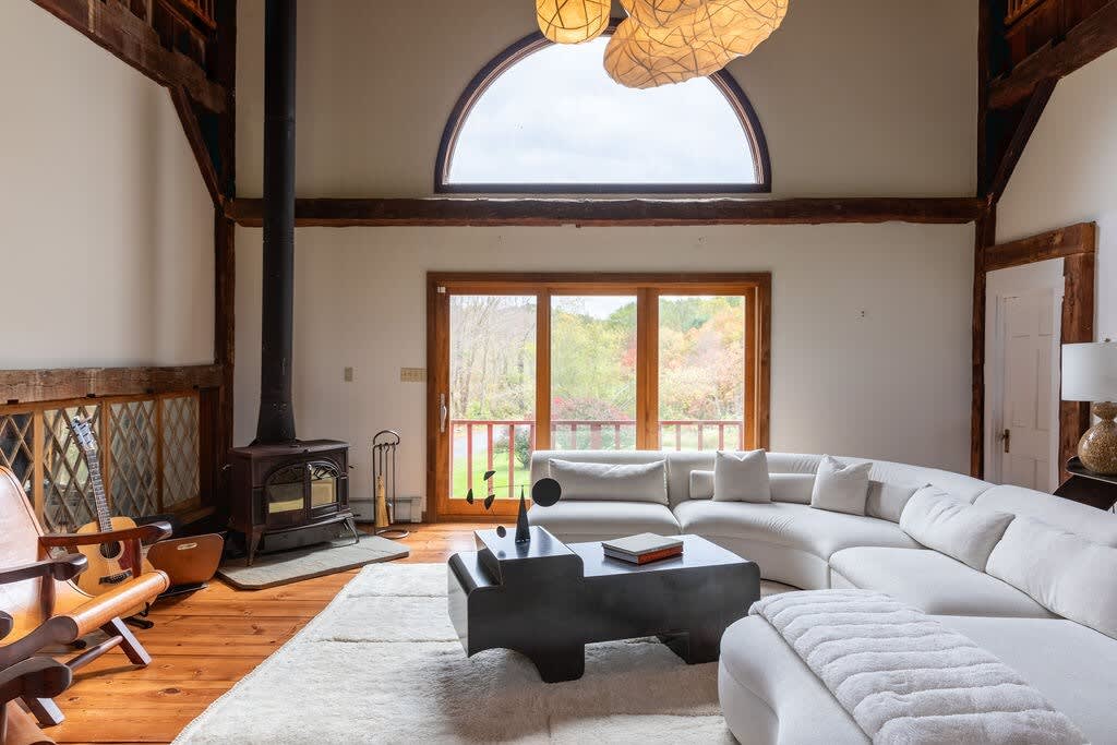 Wide shot of a living room with a white sectional sofa on a light rug, facing a dark coffee table. A black wood-burning stove is visible on the left, with a large arched window above and a sliding glass door leading to the outside, all within a rustic barn structure.