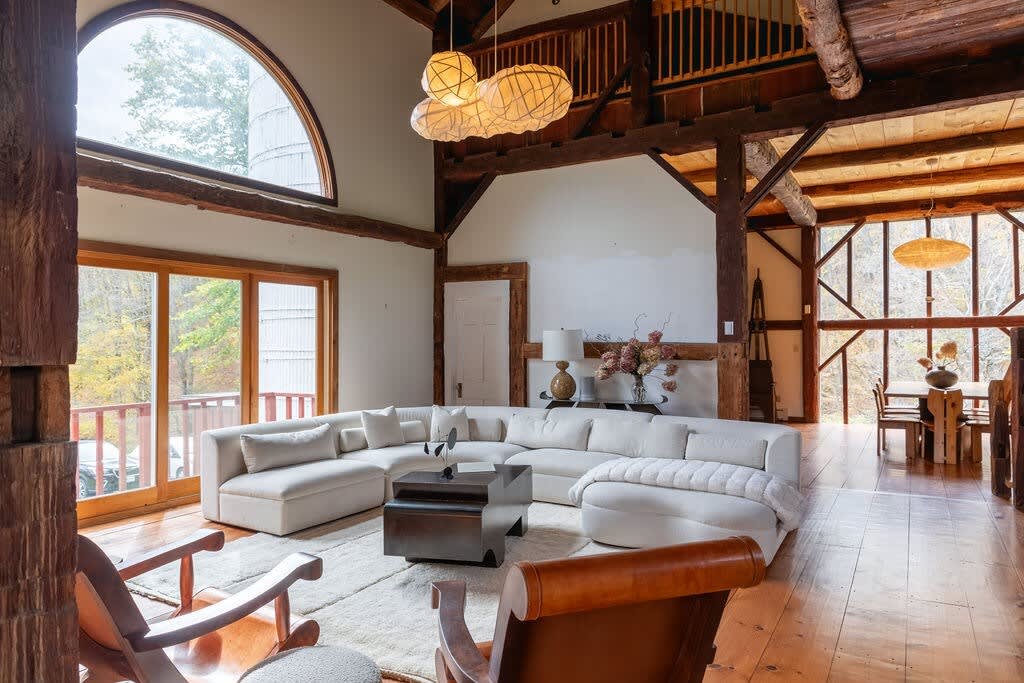 Expansive living room featuring a large white sectional sofa, exposed wooden beams, and a high vaulted ceiling. An arched window and sliding glass doors overlook the natural landscape, with a unique cloud-like light fixture overhead.