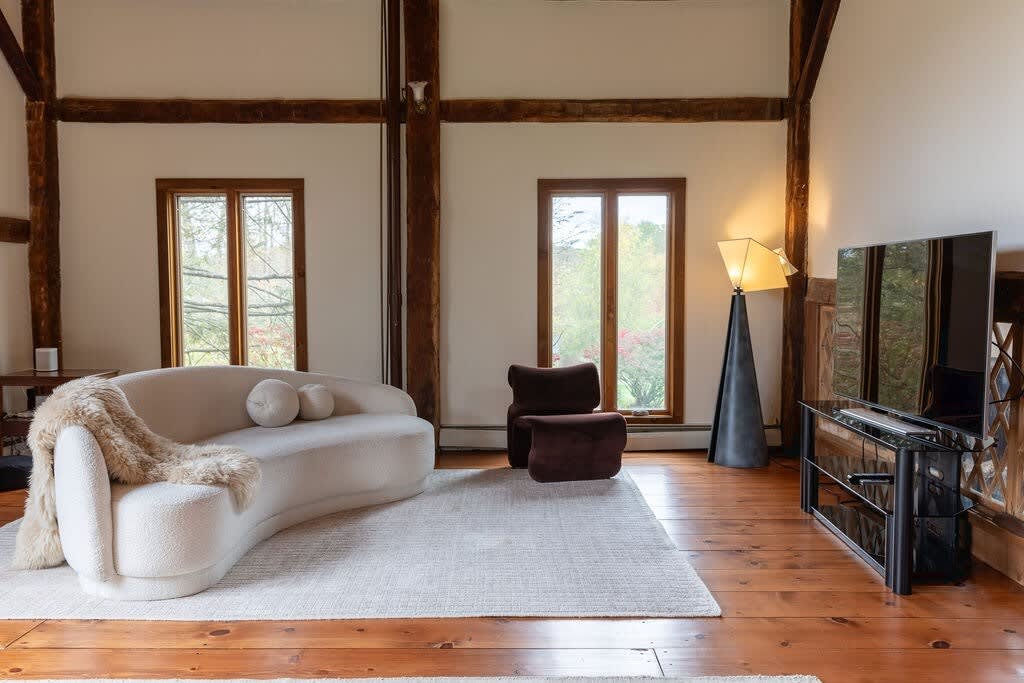 Bright living space with a curved white sofa draped with a faux fur throw, positioned on a light area rug. Two large windows offer views of greenery, and a modern floor lamp stands next to a flat-screen TV on a dark console, all on warm hardwood floors.