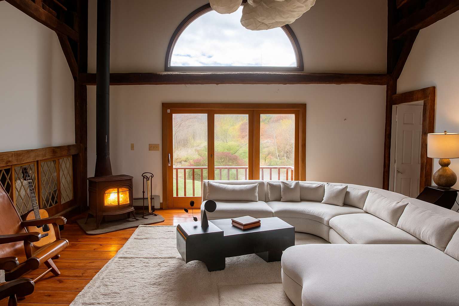 Cozy living room with a large white sectional sofa, a black coffee table, and a wood-burning stove with a fire. A large arched window and glass sliding doors offer views of autumn foliage, complemented by exposed wooden beams.