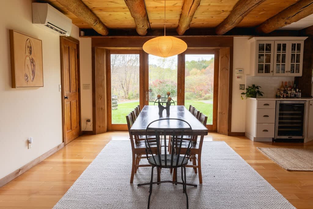 A bright dining area centered around a long wooden table with an eclectic mix of chairs, illuminated by a large paper lantern, with glass doors offering a scenic view of the outdoors.