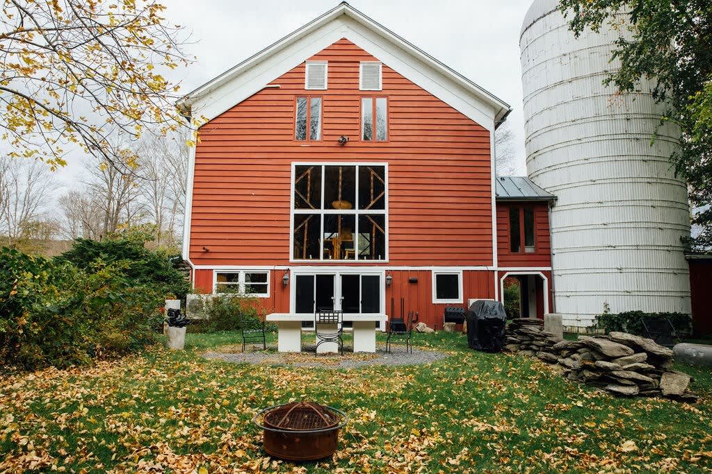 Rear view of the red barn house with a tall, multi-pane window facade and a white silo. A stone wall, outdoor seating, a BBQ grill, and a fire pit are visible in the grassy yard covered with autumn leaves.