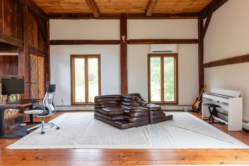 Versatile room with warm hardwood floors and a large light area rug, featuring a desk with an office chair and a white digital piano. A unique brown leather sofa adds a sculptural element, with two windows providing natural light.