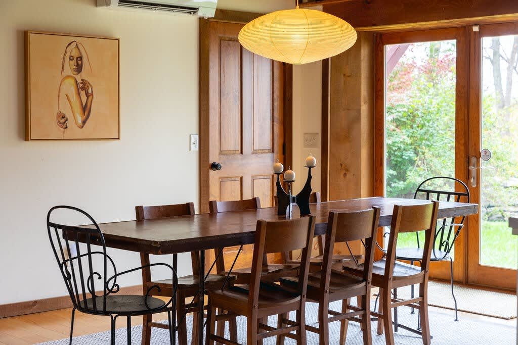 A close-up of the dining area, highlighting the rustic wooden table set with modern candle holders, various dining chairs, and a framed artwork on the wall, bathed in natural light from the glass doors.