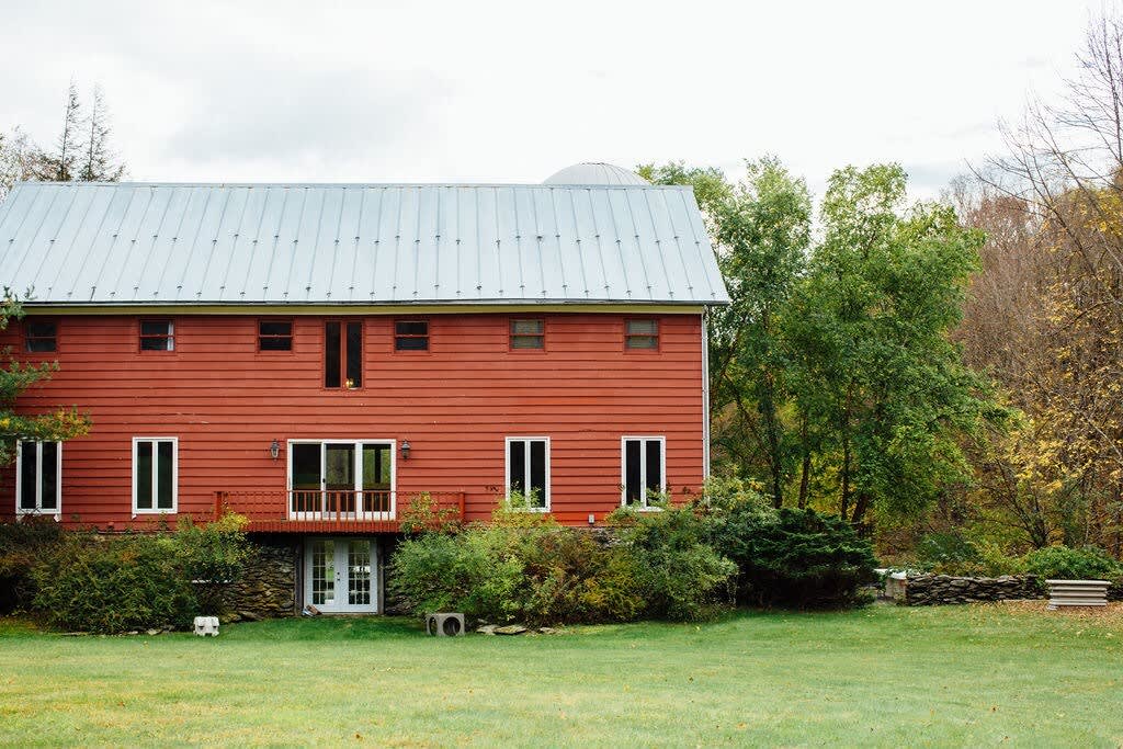 The exterior of a large red barn-style house with a silver metal roof, featuring multiple windows and a balcony, surrounded by a lush green lawn and mature trees under a cloudy sky.