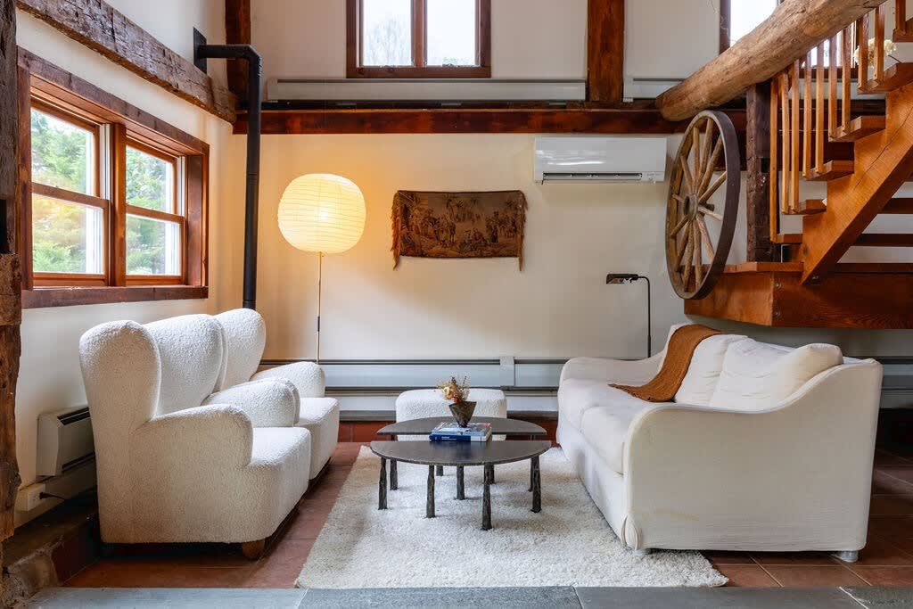 Cozy secondary living area with two white armchairs and a matching sofa, centered around a round coffee table on a plush rug. Exposed wooden beams, a large wagon wheel decor, and a wooden staircase add rustic character.