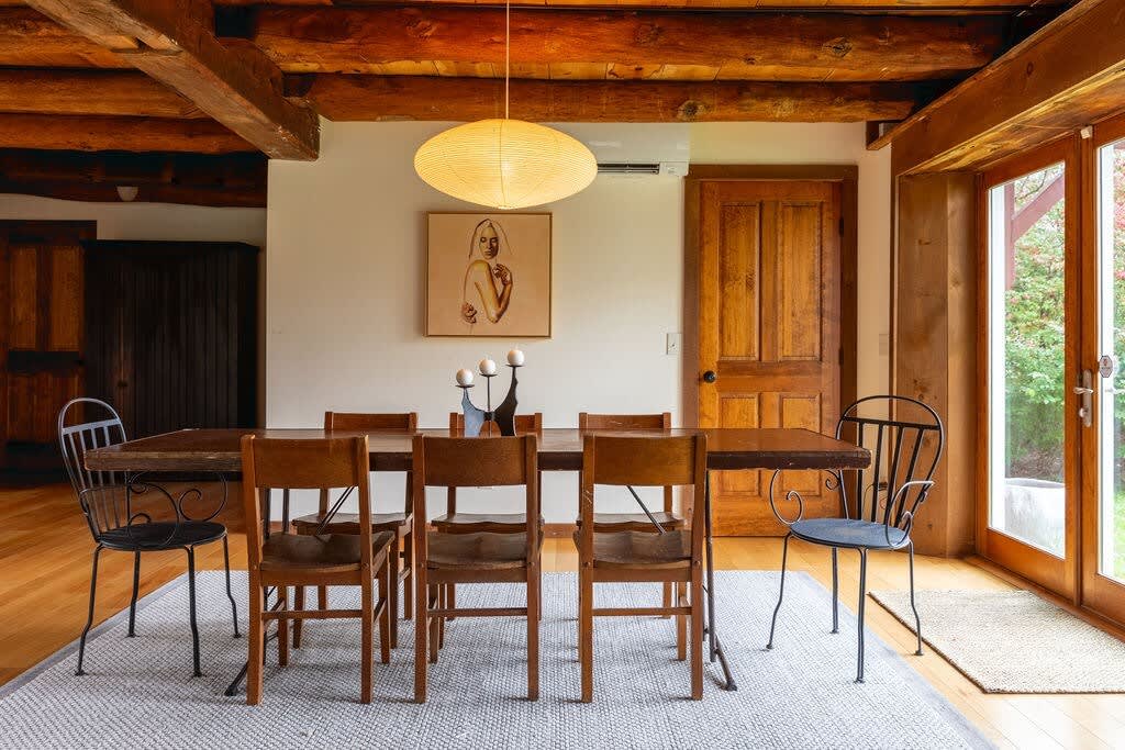 The dining area features a long wooden table with an eclectic mix of chairs, positioned under a ceiling with prominent exposed wooden beams, and large glass doors providing direct outdoor access.