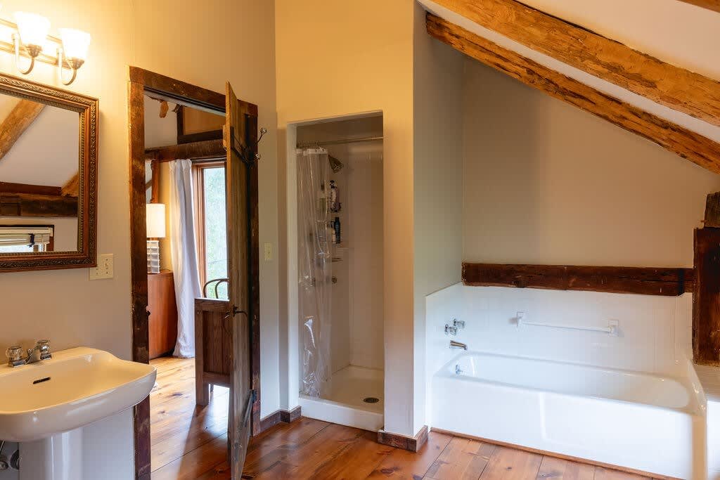 A rustic bathroom featuring a white pedestal sink, a separate shower stall with a clear curtain, and a white bathtub, all set against warm walls and rich hardwood floors with exposed wooden beams overhead.