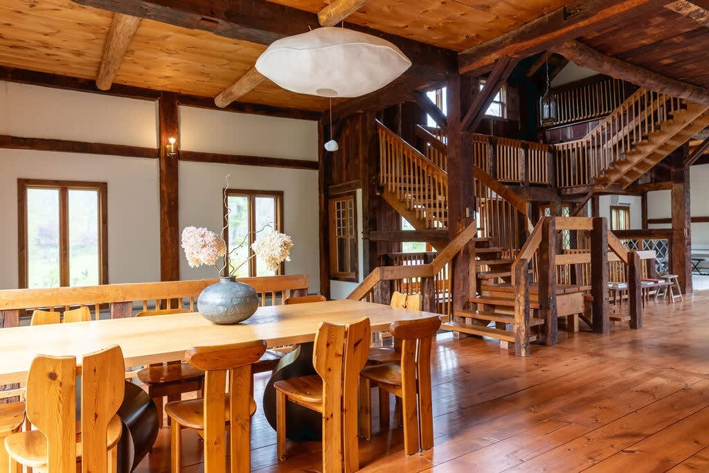 Wide view of the dining area featuring a long wooden table with matching chairs and a bench, set on polished hardwood floors. A grand wooden staircase with intricate railings is visible in the background, complementing the exposed wooden beams throughout the space.