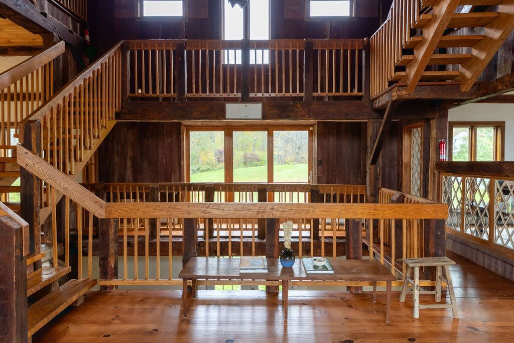 A grand multi-level interior space with exposed wooden beams, intricate wooden staircases, and railings, featuring a central wooden table and a view of the outdoors through large windows.