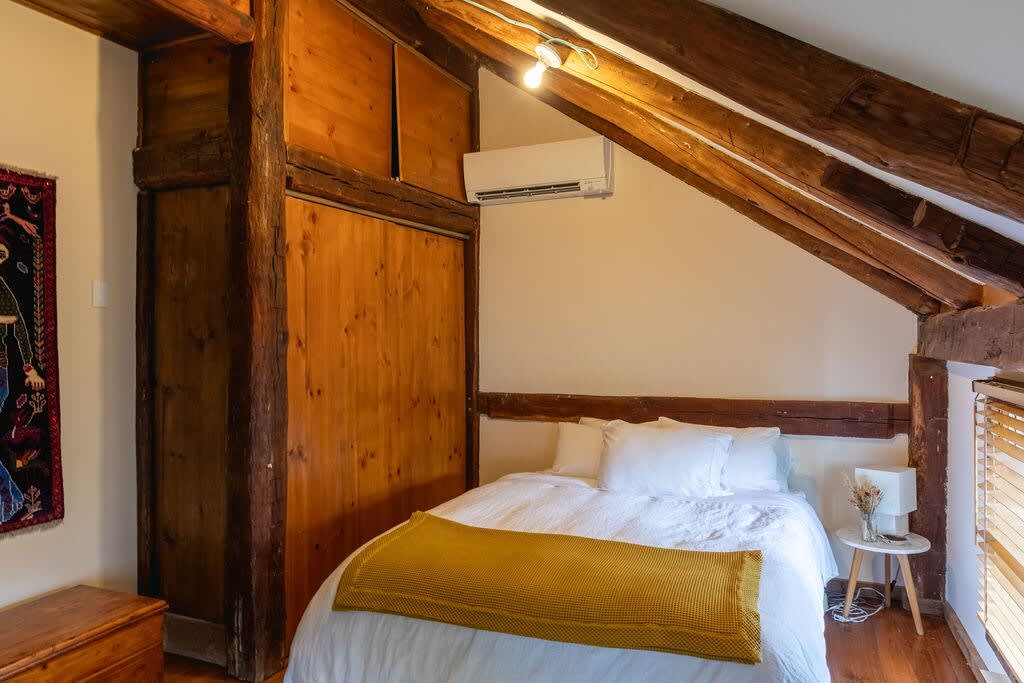 A cozy bedroom with a white bed and yellow throw, featuring sloped ceilings with exposed wooden beams, built-in wooden storage, and an air conditioning unit mounted on the wall.