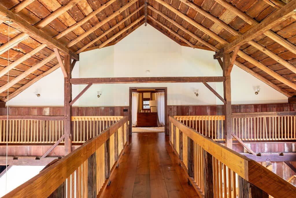 A stunning interior view of a renovated barn, showcasing a wooden bridge or walkway with rustic railings, leading towards a distant room, all beneath a high vaulted ceiling with intricate exposed wooden beams.