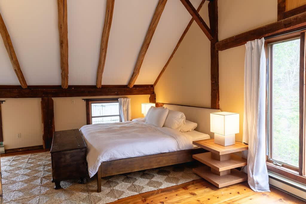 Cozy bedroom with a queen-sized bed featuring white bedding and a light headboard, set on a patterned area rug over hardwood floors. Exposed wooden beams define the vaulted ceiling, and a wooden chest sits at the foot of the bed, with a large window providing natural light.