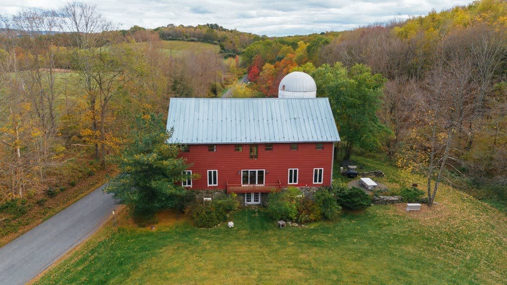 An aerial view of the red barn-style house with a silver roof, situated on a green hill surrounded by vibrant autumn foliage and a winding road, showcasing the expansive rural landscape.