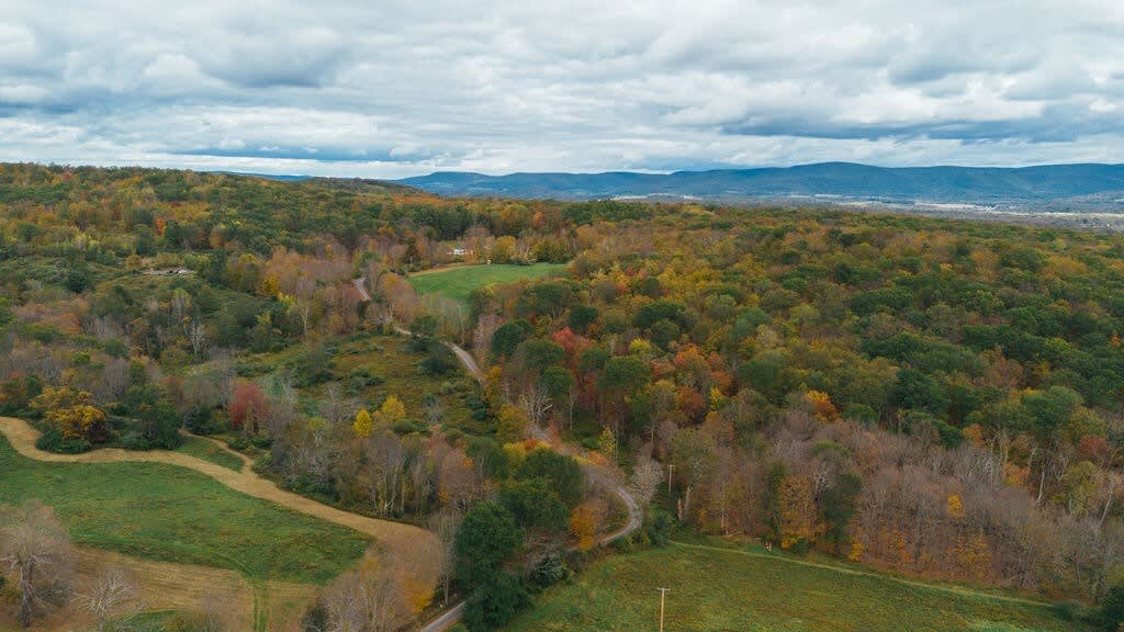 Wide aerial shot showcasing the property's vast natural surroundings, with rolling hills covered in dense autumn forests and a winding road. The barn house is nestled within this picturesque, secluded landscape.