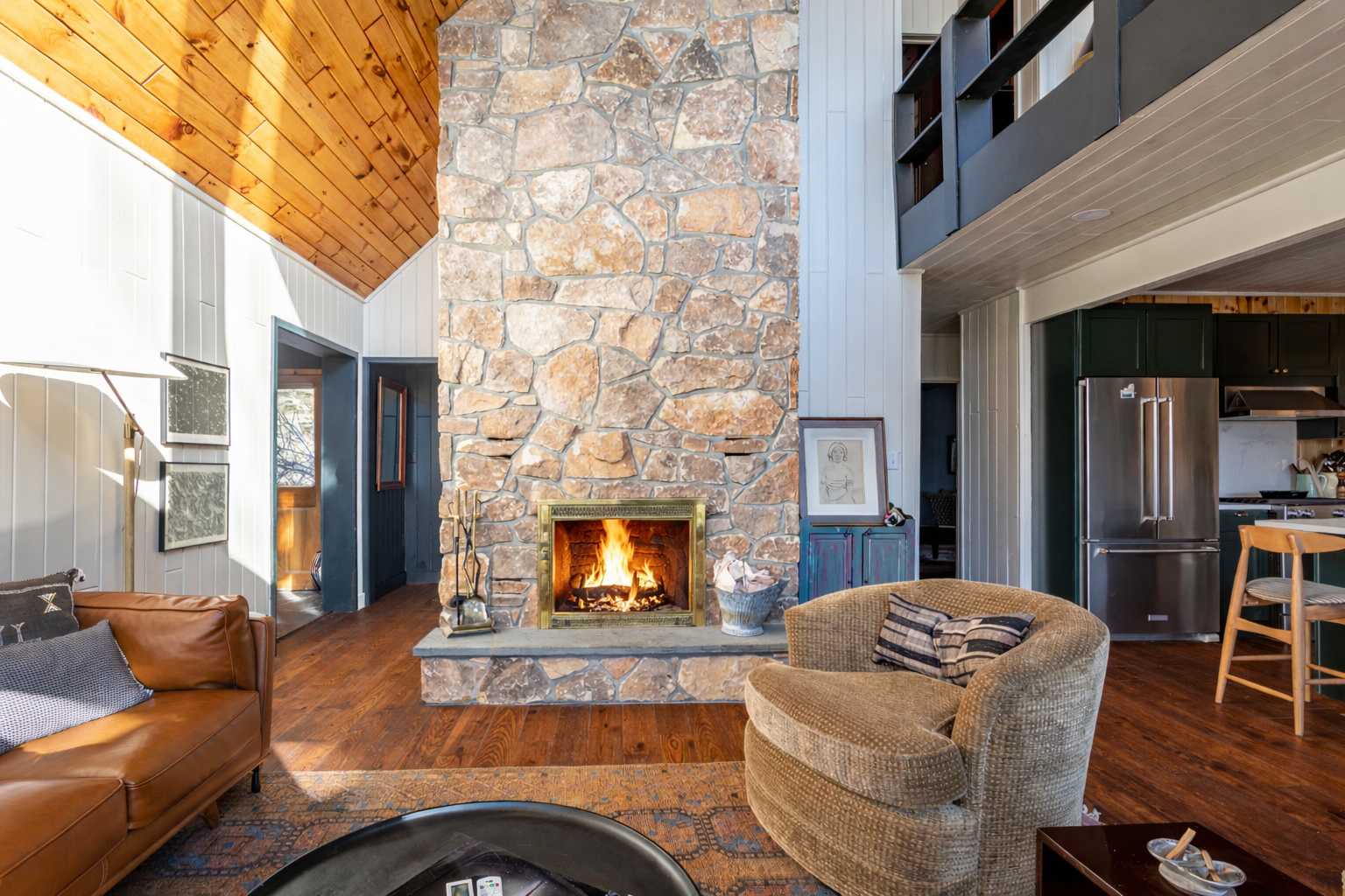 Spacious living room centered around a tall stone fireplace, featuring a brown leather sofa, a plush armchair, and a view towards a modern kitchen with dark green cabinets.