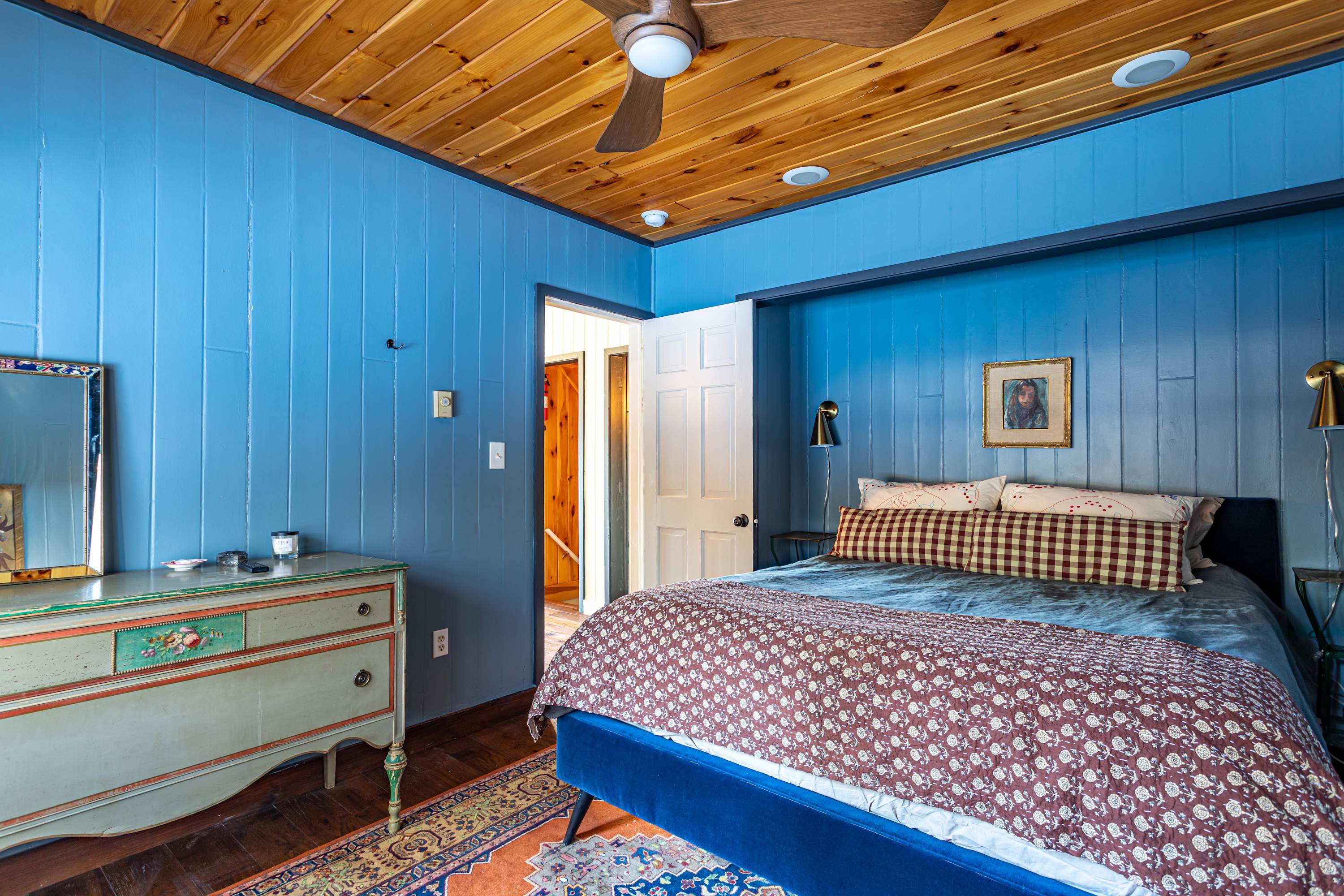 View of the blue-walled bedroom showing a bed with a dark blue frame and patterned quilt, a vintage-style dresser with a mirror, and a wooden ceiling with a fan.