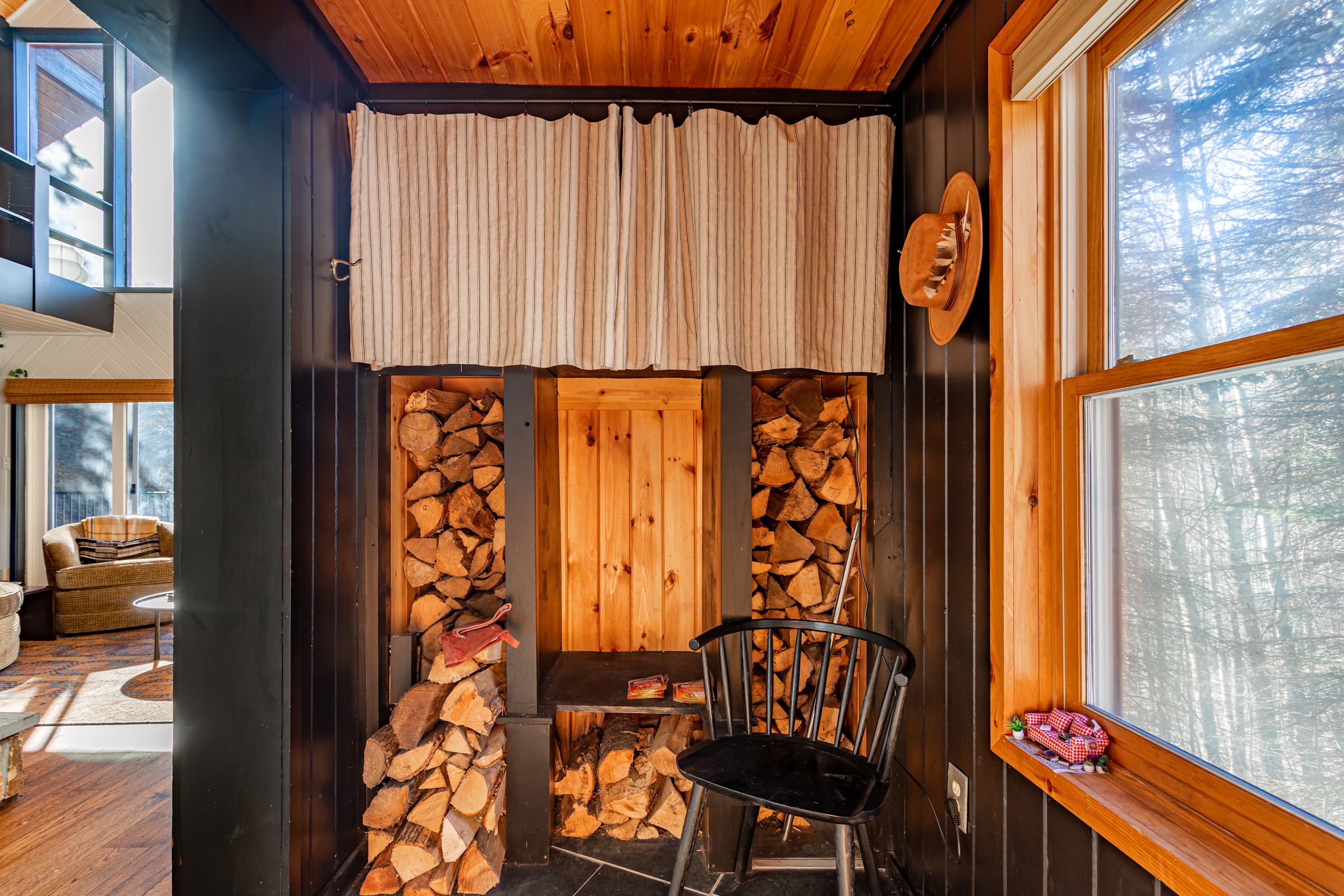 Rustic entry area or mudroom with stacked firewood storage, a small black chair, and a window, featuring dark wood paneling and a natural wood ceiling.