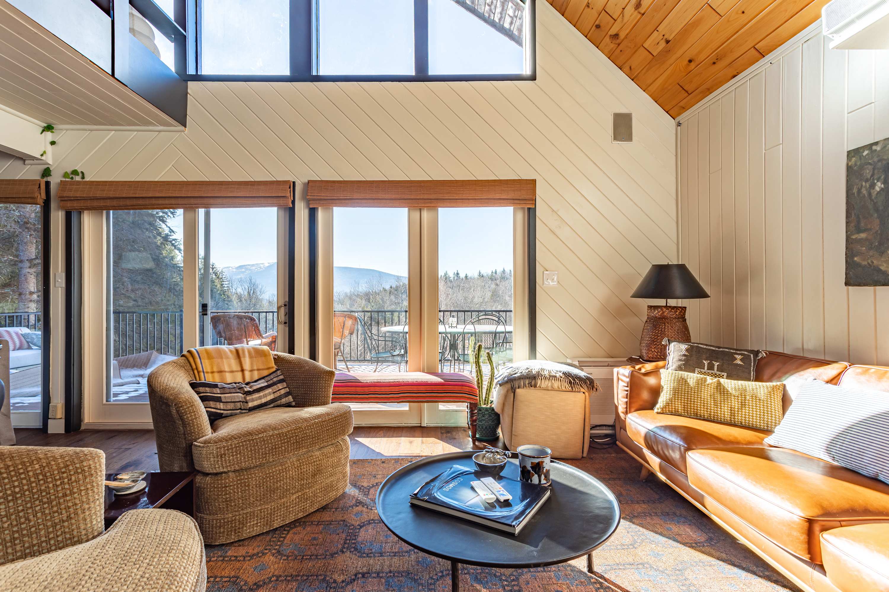 Bright living room with a vaulted wooden ceiling, light-colored paneled walls, and large windows offering mountain views, furnished with a brown leather sofa and a patterned rug.