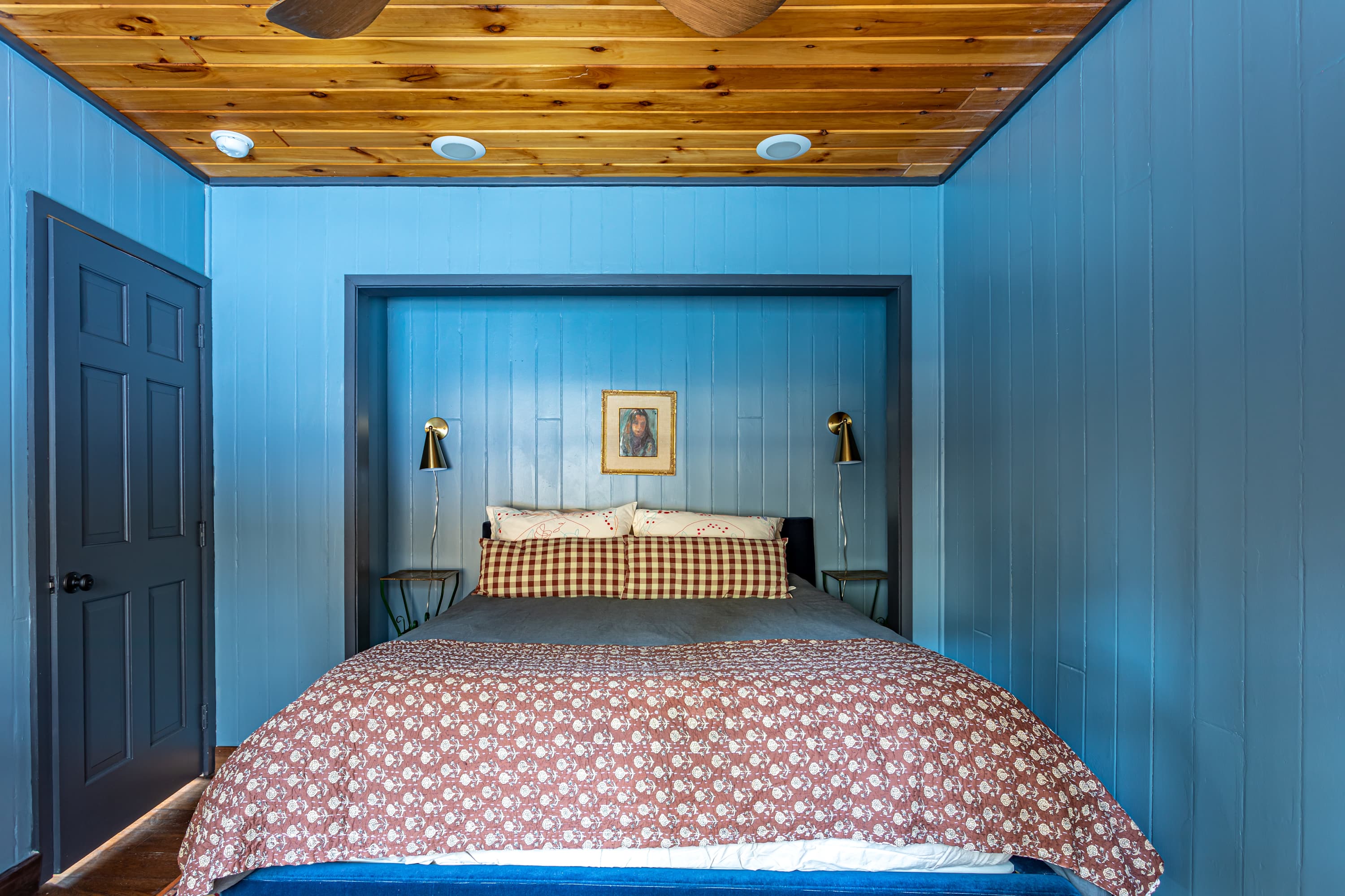 Serene bedroom with blue paneled walls, a wooden ceiling, and a bed recessed into an alcove with brass sconces, featuring a patterned quilt and a dark blue bed frame.