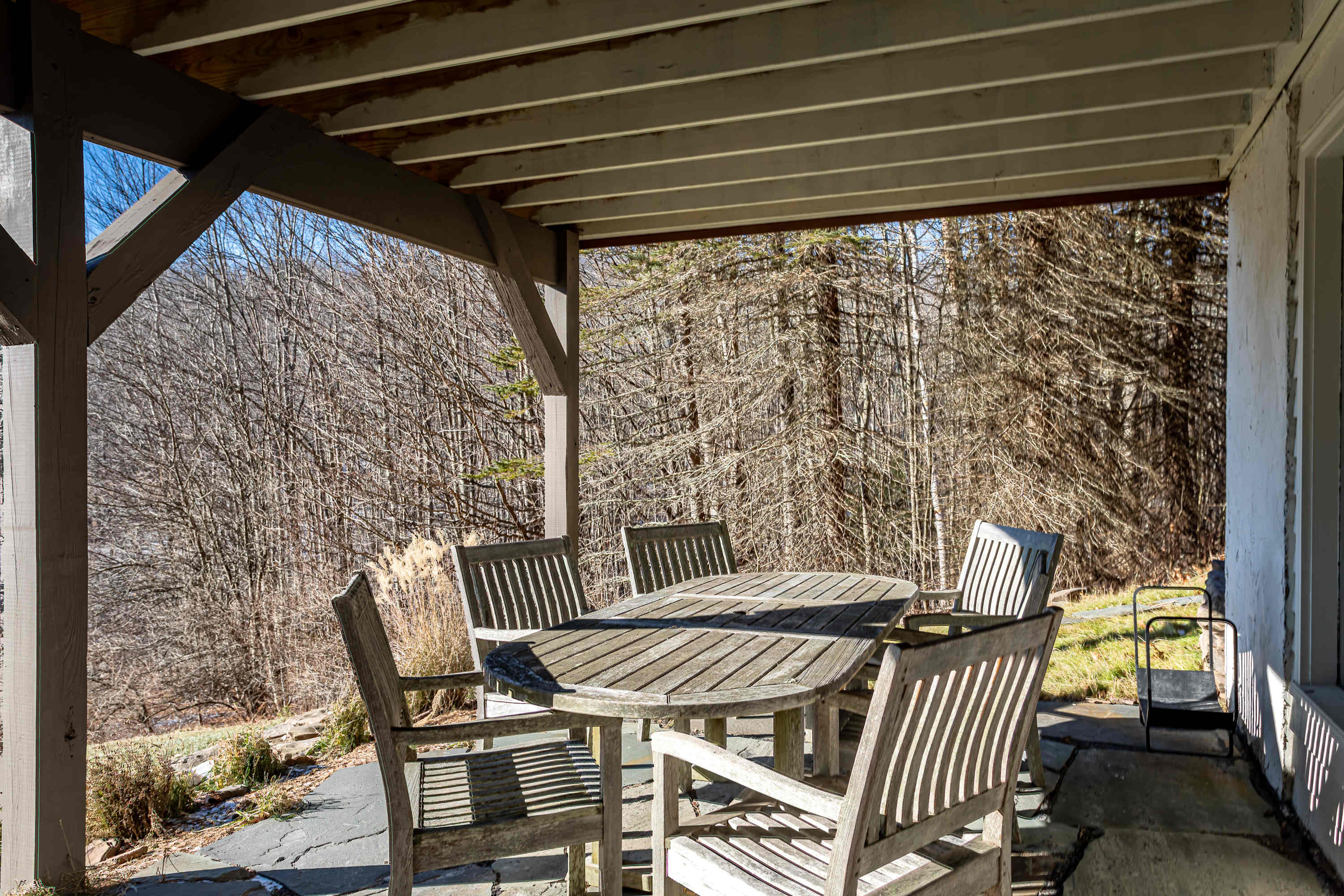 Shaded outdoor deck area featuring a round wooden dining table and matching chairs, set against a backdrop of bare winter trees. The deck is covered by an overhead structure, providing a comfortable outdoor seating space.