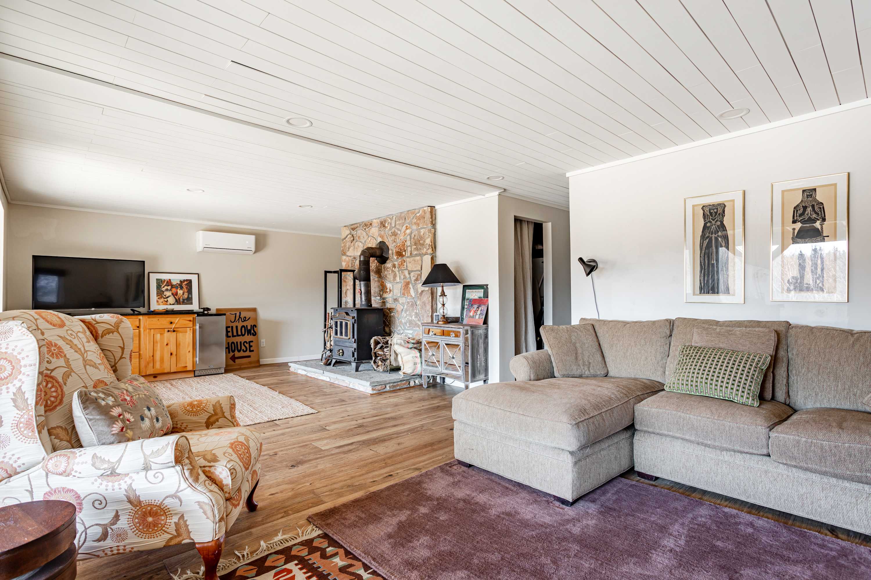 Expansive living room showcasing a rustic wood-burning stove on a stone hearth, a comfortable sectional sofa, and a patterned armchair. The space features light wood floors, a shiplap ceiling, and ample natural light from large windows.