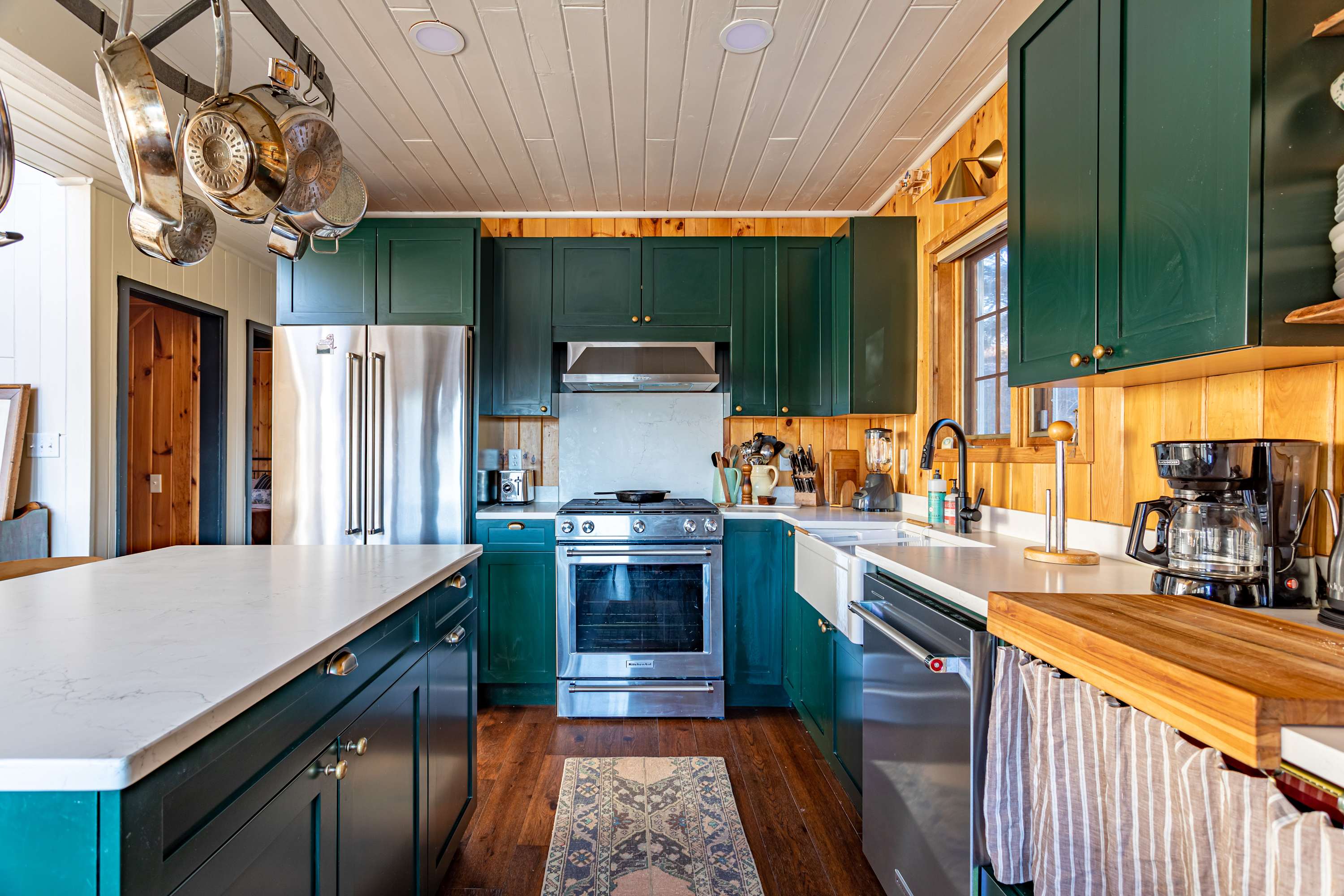 Modern kitchen featuring dark green cabinetry, stainless steel appliances, a white farmhouse sink, and natural wood paneled walls, with a patterned rug on the hardwood floor.