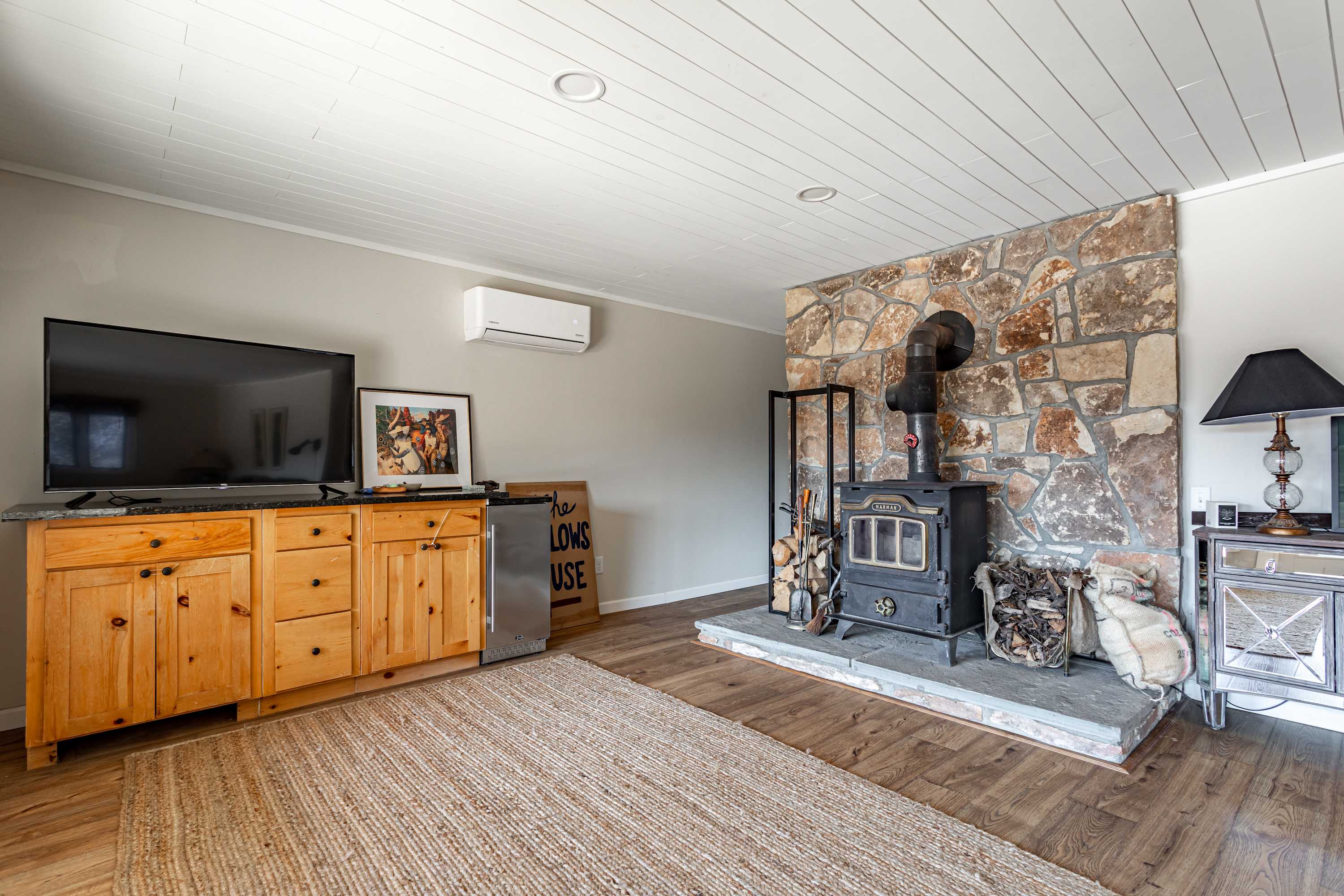Inviting living room with a prominent wood-burning stove set against a rustic stone wall, complemented by a wooden TV console and a side table with a lamp. A natural fiber rug grounds the space, with a mini-split AC unit on the wall.