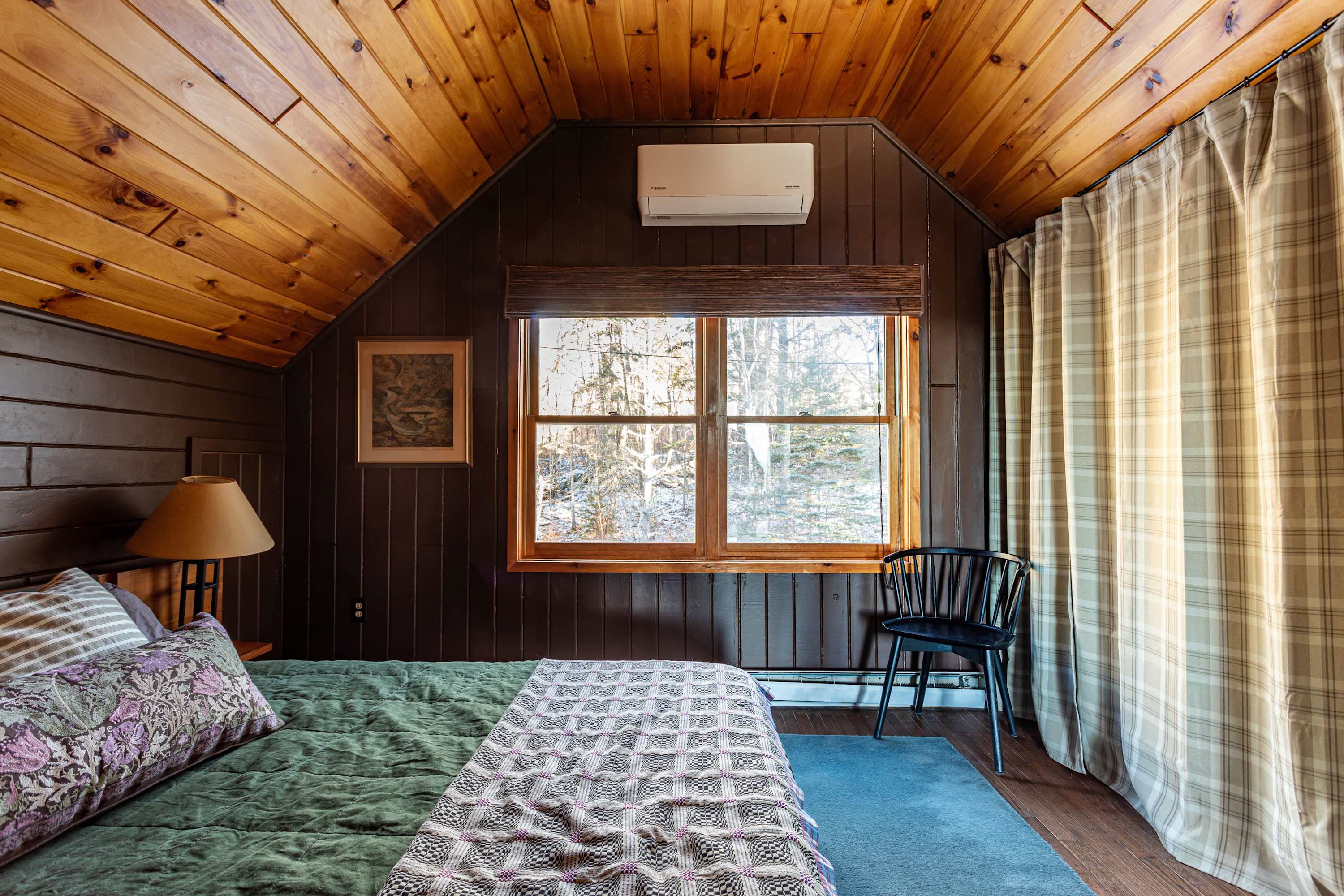 A cozy bedroom with dark wood paneling and a sloped wood-paneled ceiling, featuring a bed with green and patterned bedding, a window overlooking a snowy landscape, and a wall-mounted AC unit.
