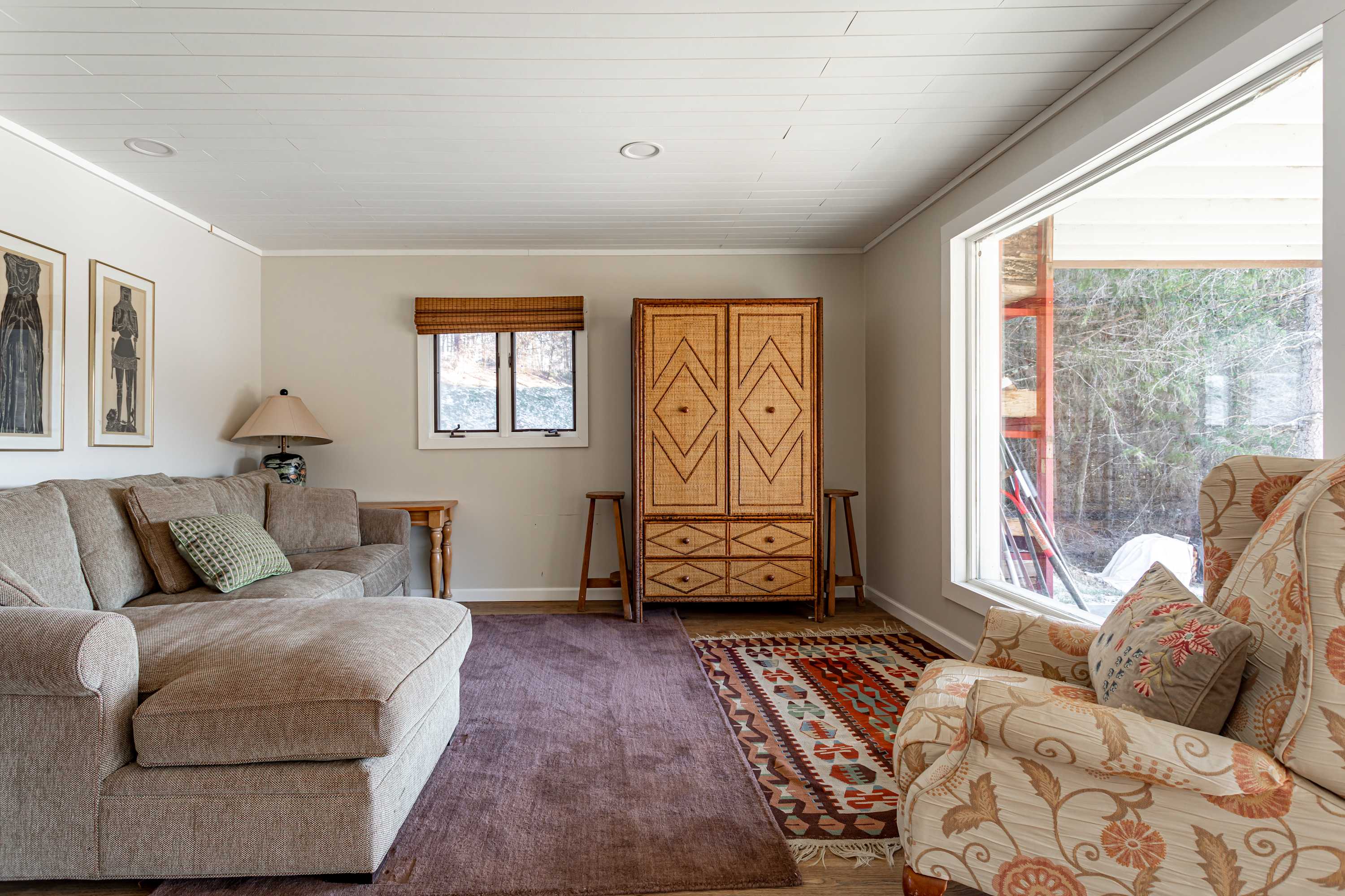 Relaxing living room corner with a plush sectional sofa, a floral-patterned armchair, and a unique woven cabinet. A window with a bamboo blind overlooks the wooded surroundings, with a colorful area rug on the light wood floor.