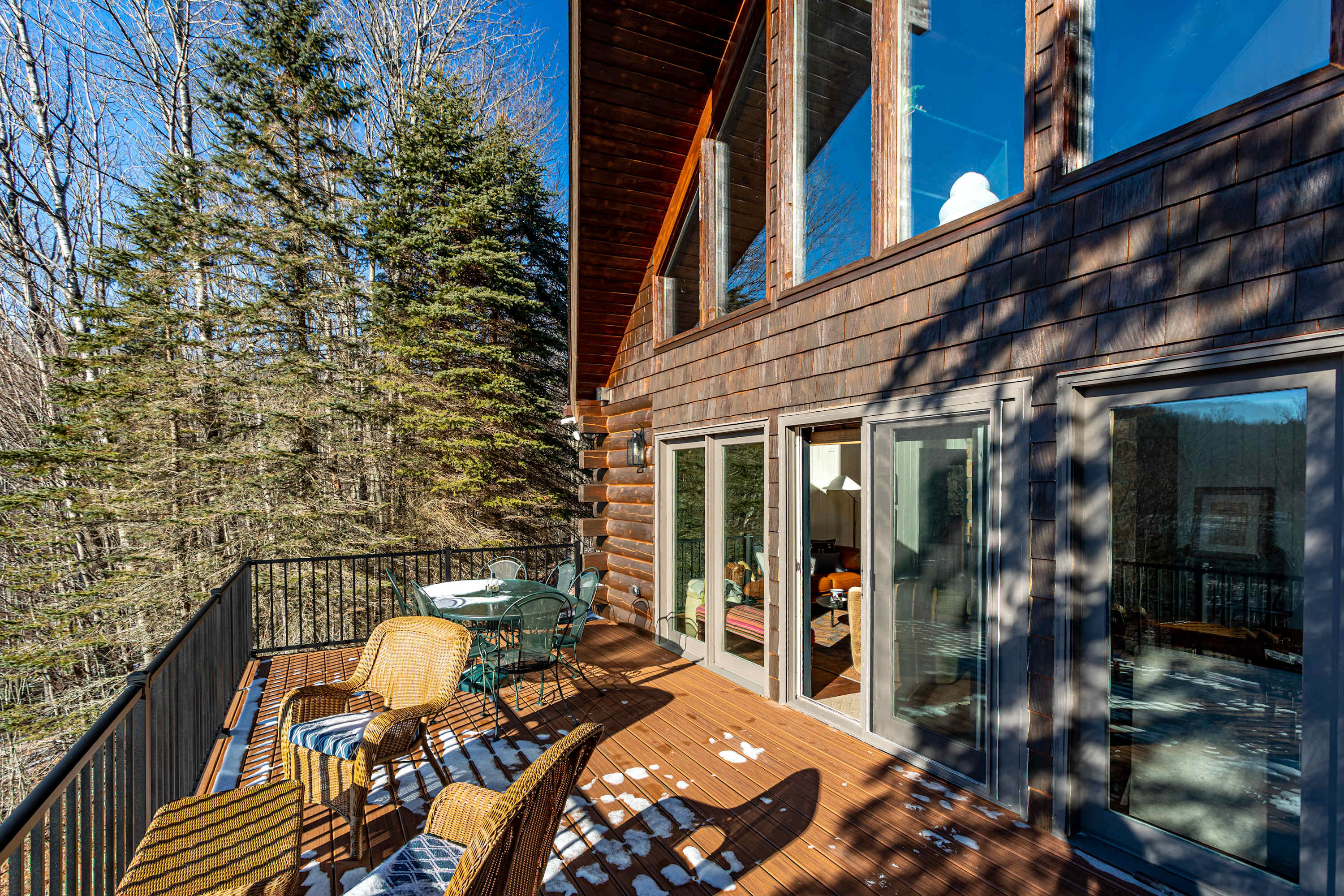 A spacious outdoor deck with a metal railing, featuring a glass-top table with chairs and wicker seating, surrounded by tall pine trees and offering glimpses of a snowy landscape.
