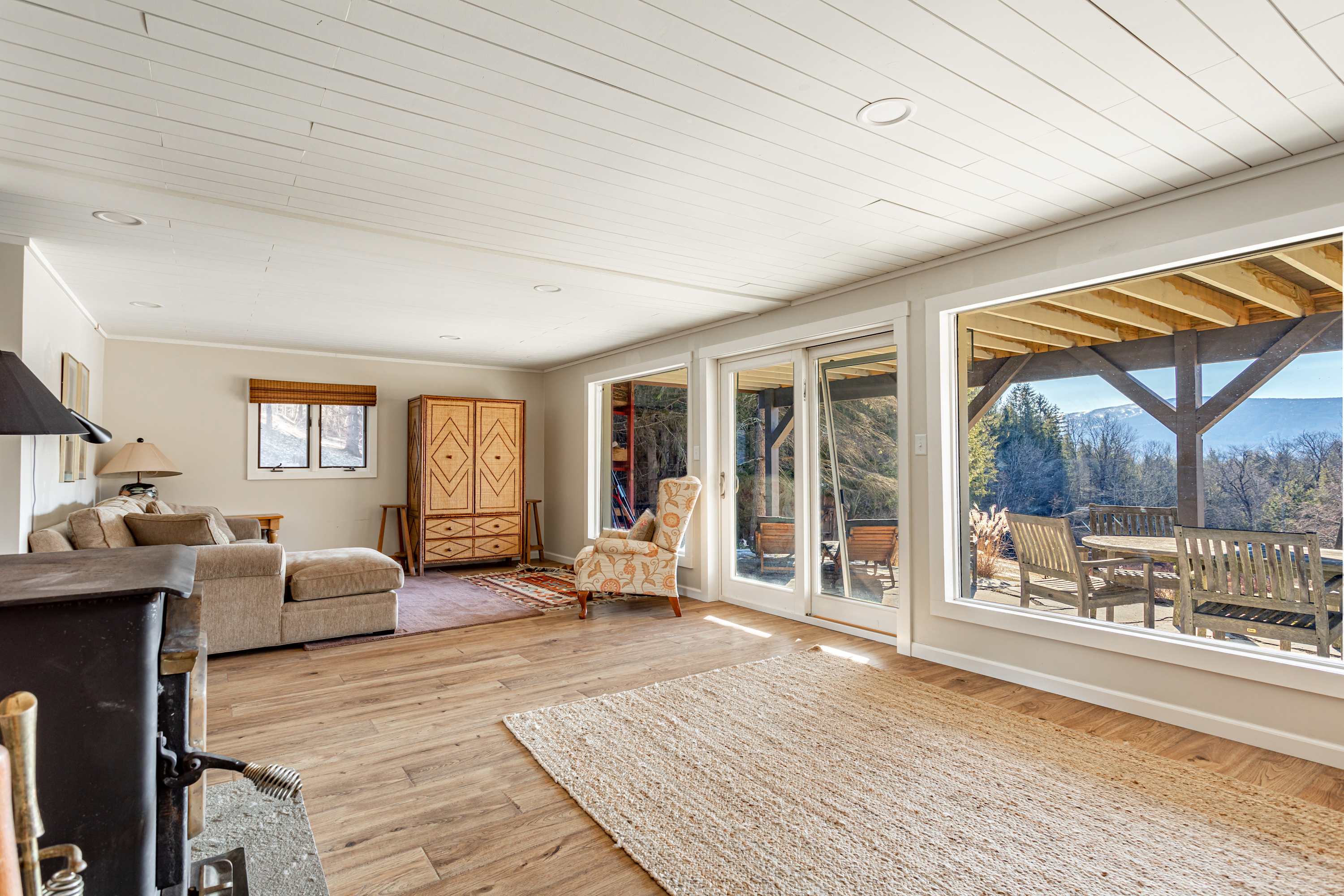 Bright living room with a comfortable sectional sofa, a patterned armchair, and expansive sliding glass doors that open to a deck with stunning mountain and forest views. Light wood floors and a shiplap ceiling enhance the airy feel.