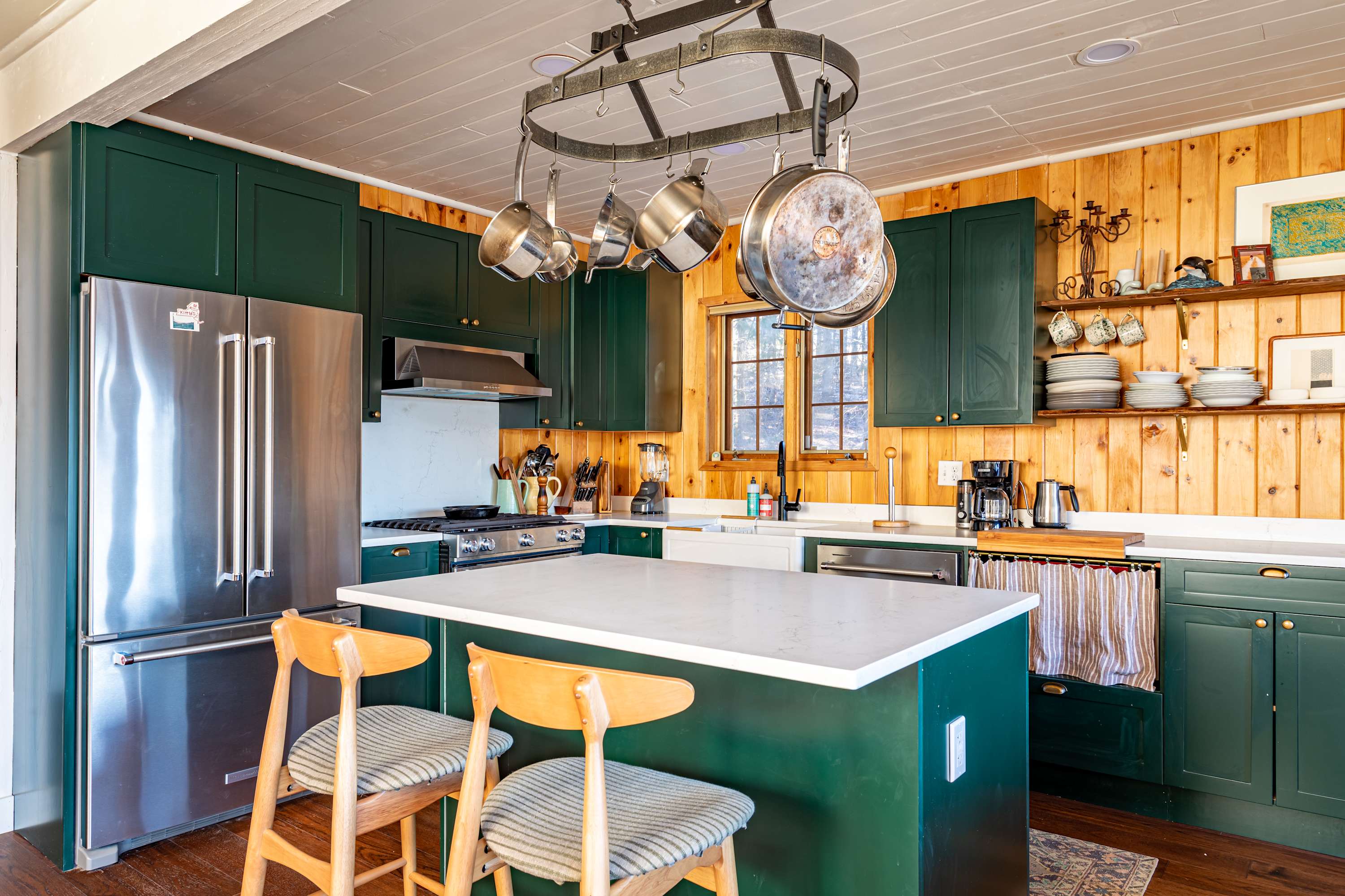 Wide view of the kitchen with dark green cabinetry, a large white island with seating, stainless steel appliances, and natural wood paneled walls, illuminated by a window.