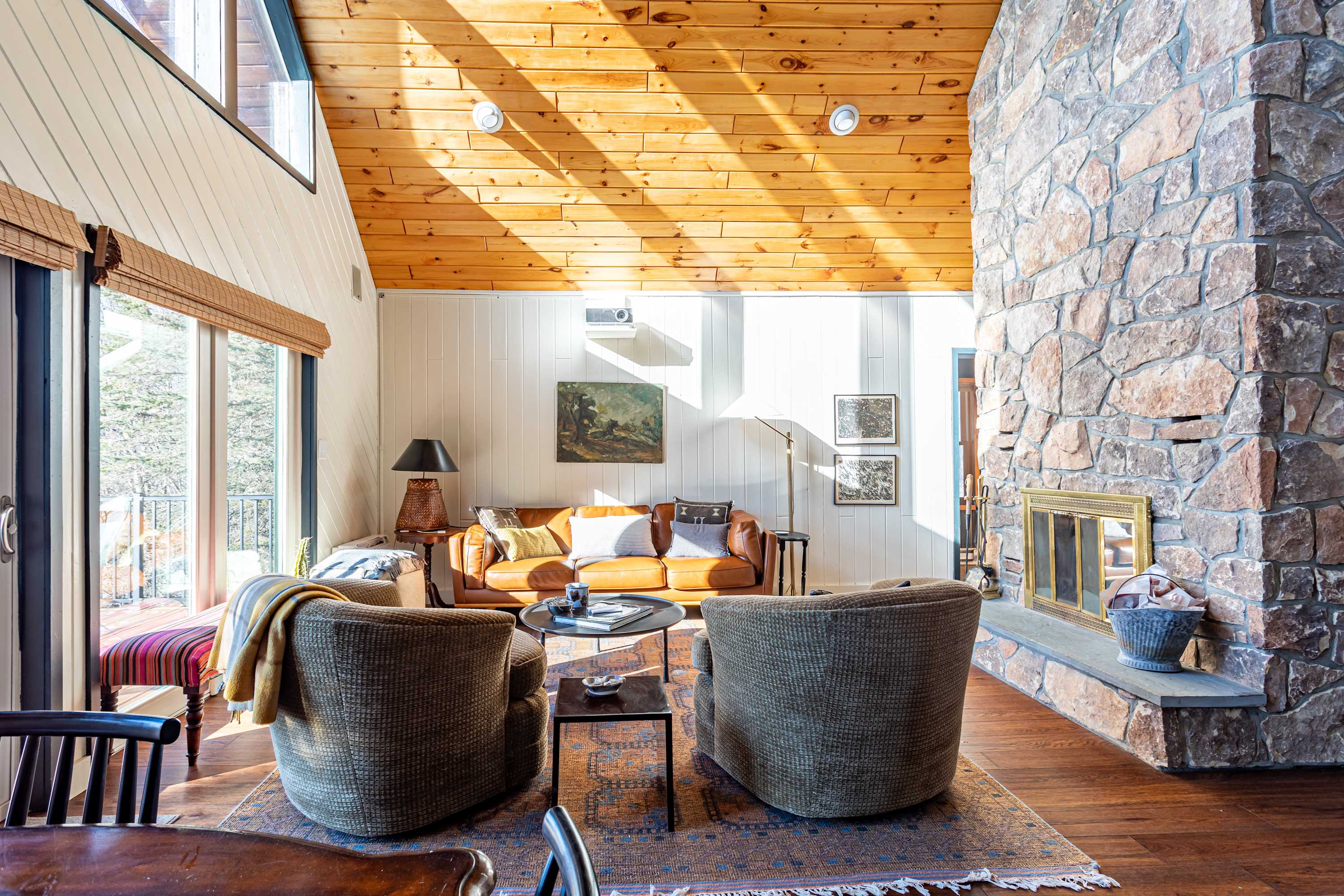 Cozy living room with a large stone fireplace, a vaulted wooden ceiling, and a brown leather sofa, featuring a patterned rug and natural light streaming through windows.