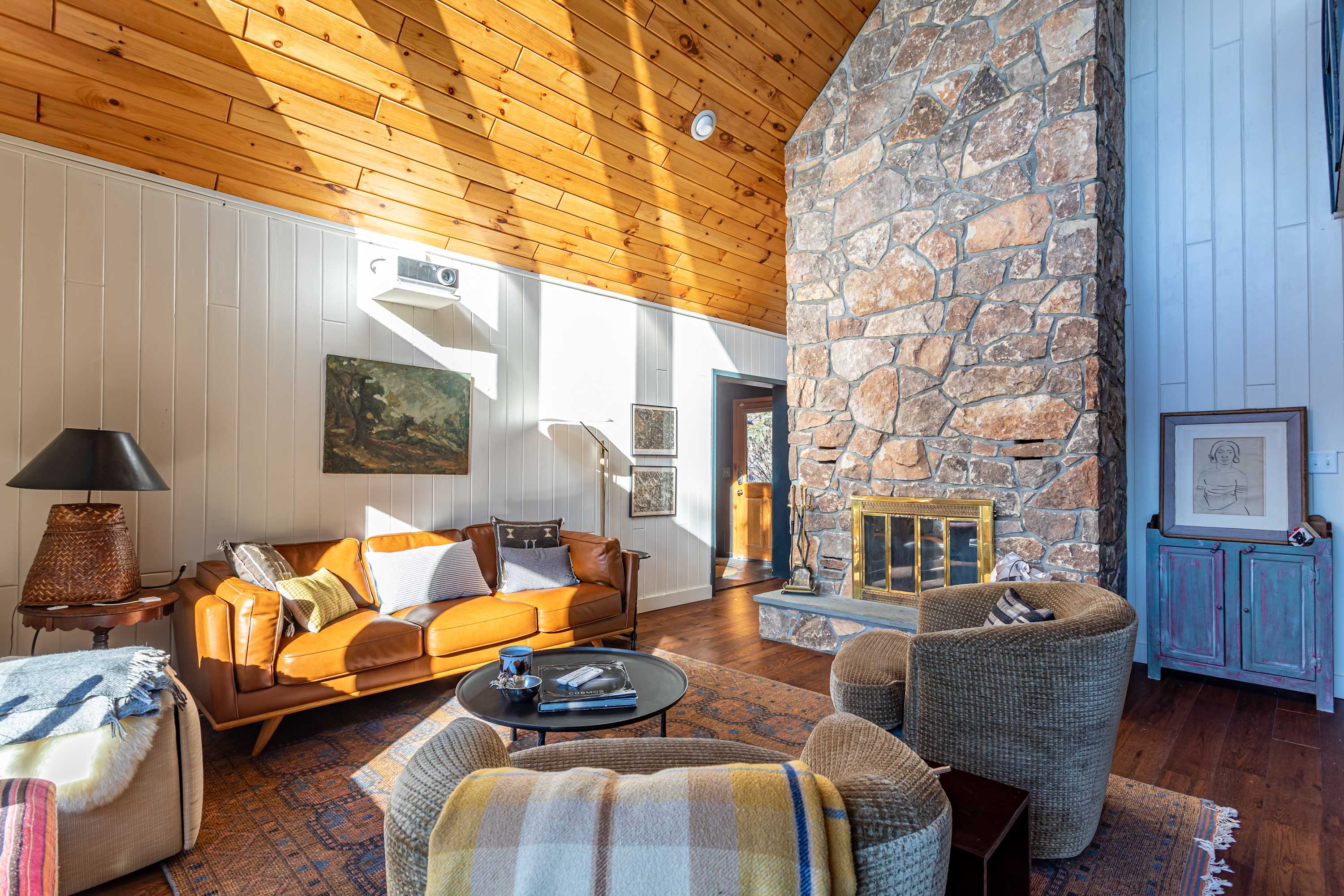 Sun-drenched living room with a tall stone fireplace, a brown leather sofa, and two armchairs on a patterned rug, featuring vaulted wooden ceilings and white paneled walls.