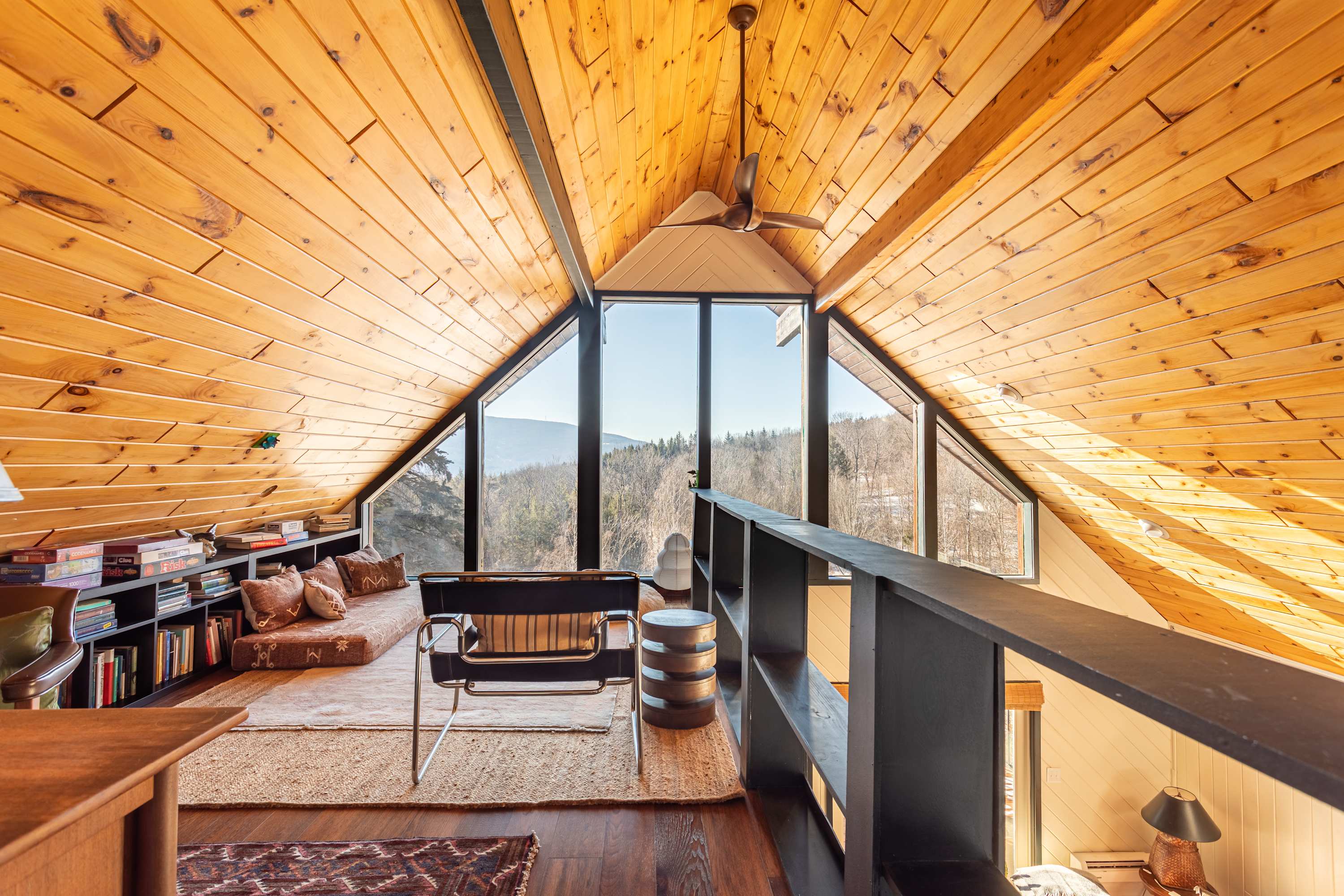 Bright loft space with sloped wooden ceilings and large triangular windows offering panoramic mountain views, furnished with a low daybed, a modern chair, and bookshelves.