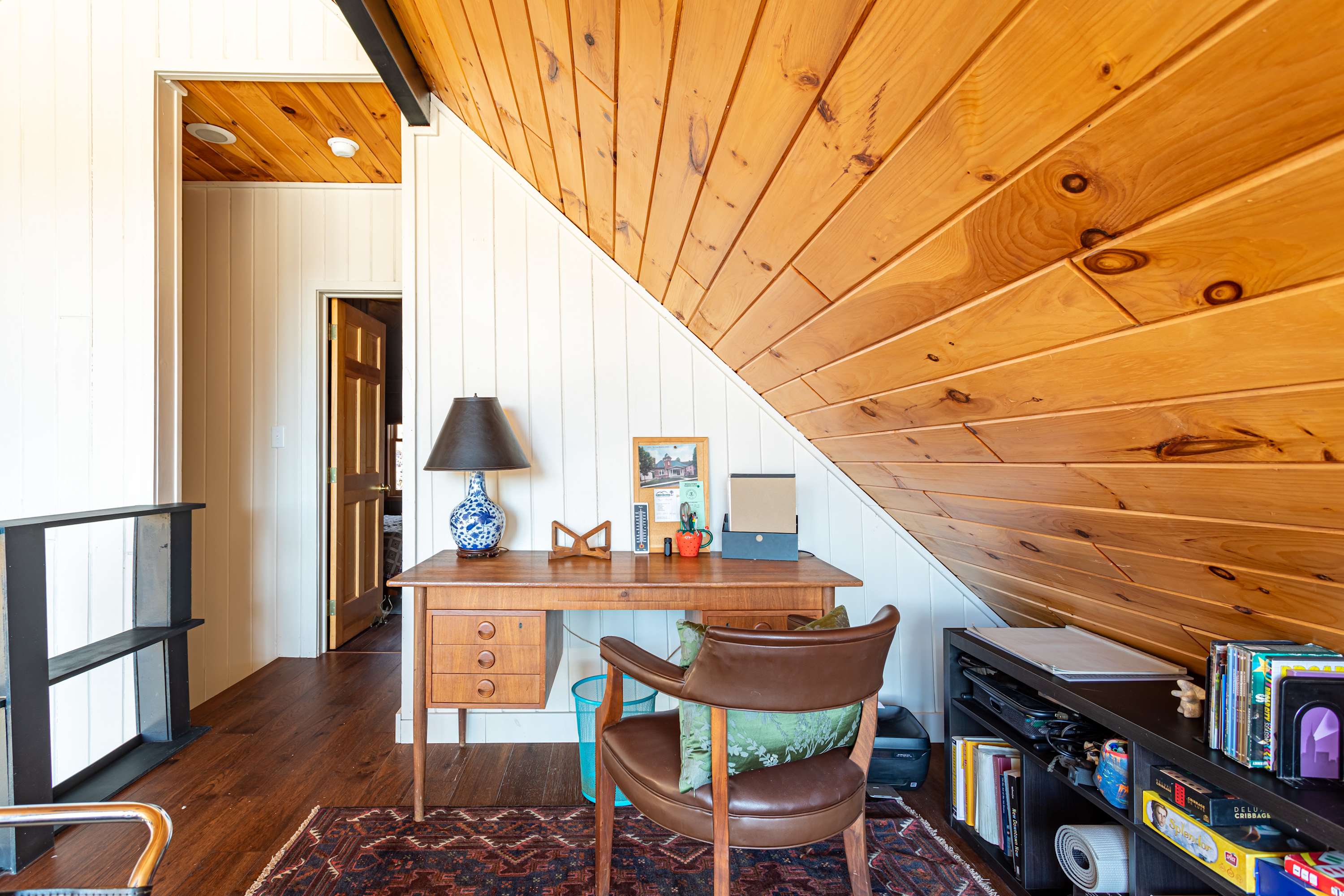 A bright office space with a sloped wood-paneled ceiling and white walls, featuring a wooden desk with a lamp, a leather chair, a bookshelf, and a view over a railing.