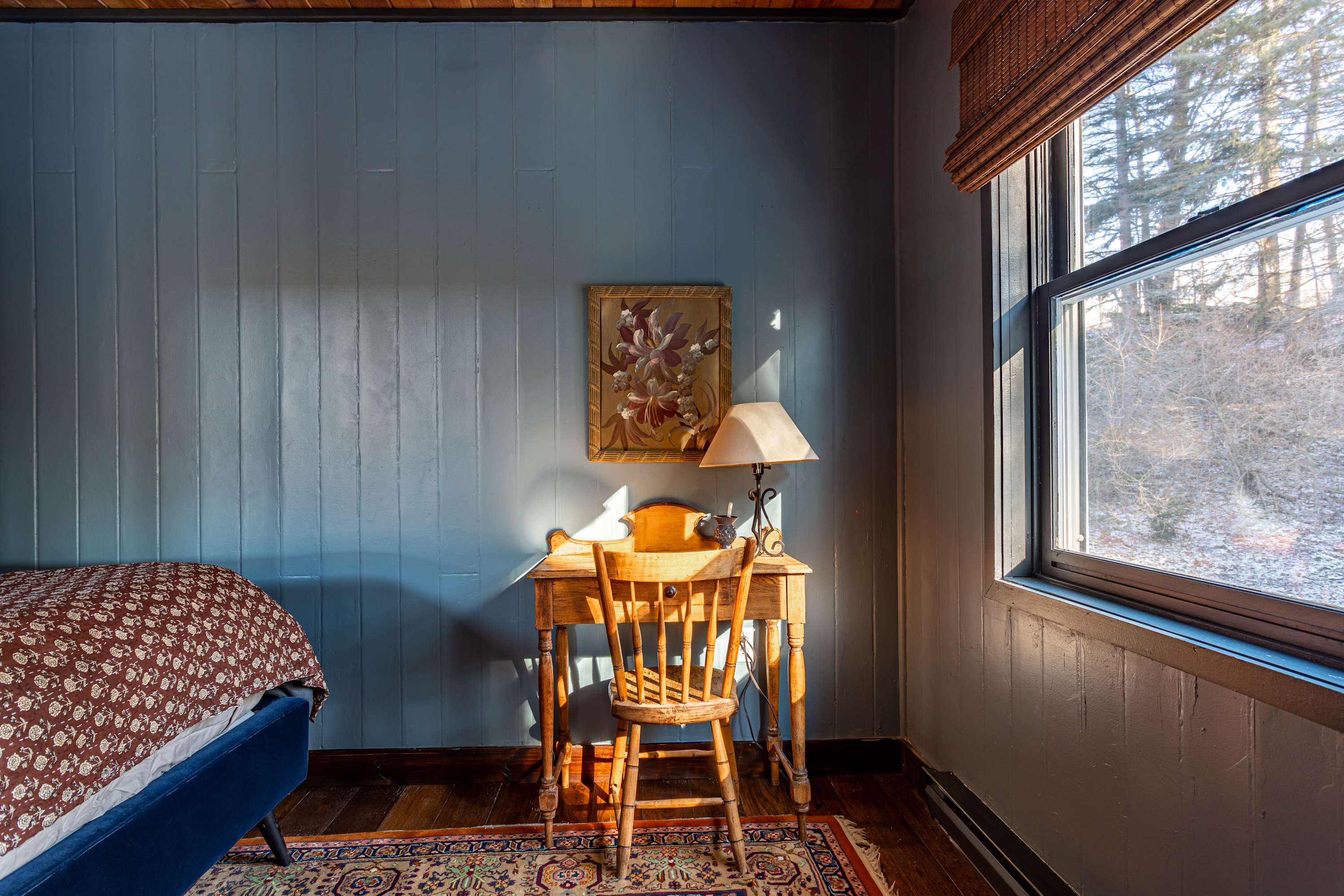Bright corner of a blue-walled bedroom featuring a wooden desk and chair next to a large window, with a painting on the wall and a patterned rug on the hardwood floor.