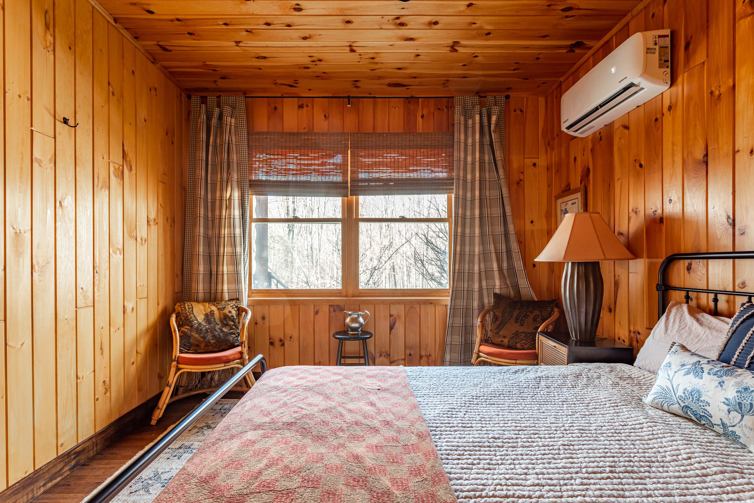 Inviting bedroom with natural wood paneled walls and ceiling, a black metal bed frame with a white and red patterned quilt, and two wicker chairs by a window overlooking trees.