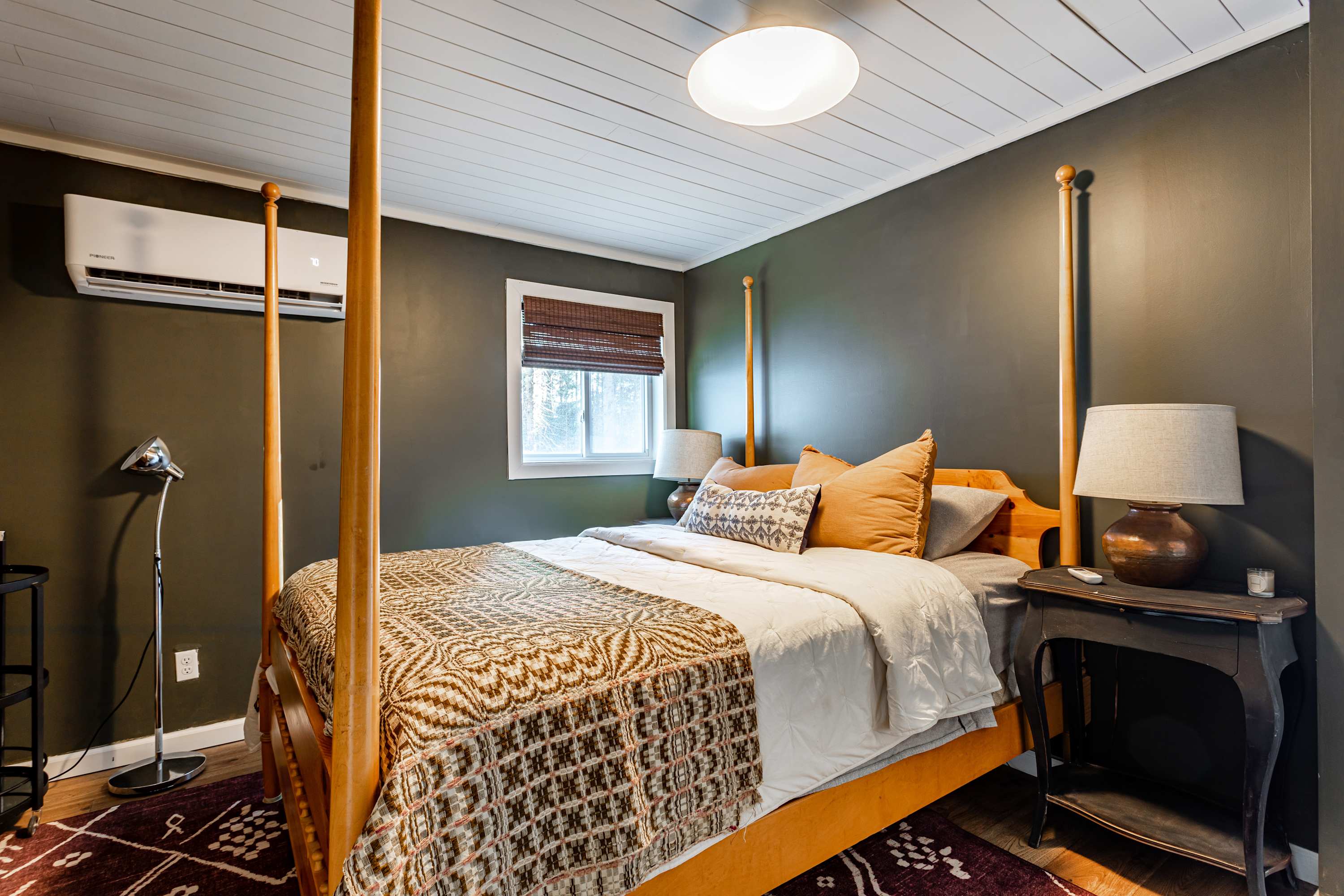 A serene bedroom with dark green walls and a white paneled ceiling, featuring a wooden four-poster bed with neutral bedding, two nightstands with lamps, and a wall-mounted AC unit.