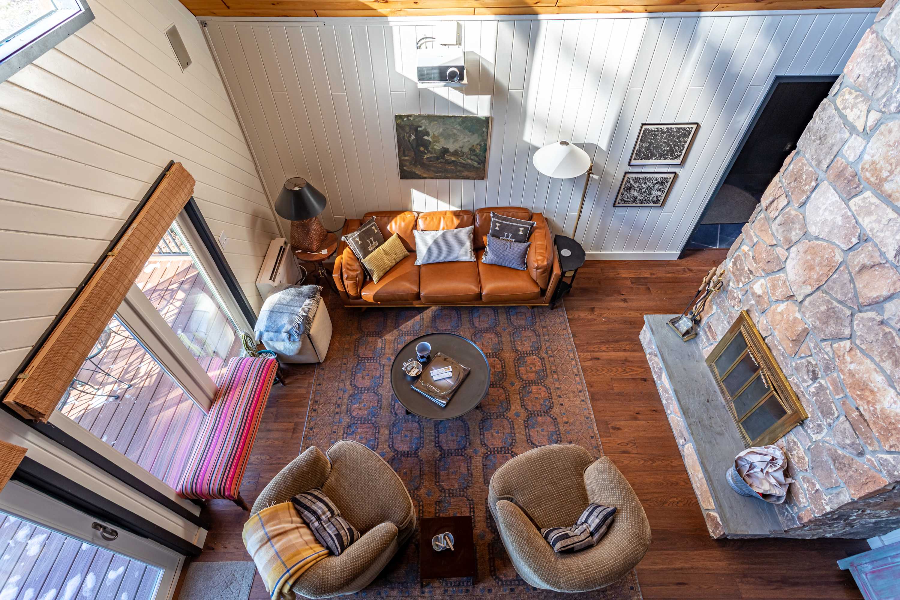 Overhead view of a sunlit living room with a brown leather sofa, two armchairs, and a round coffee table on an ornate rug, facing a large stone fireplace and a projector screen.