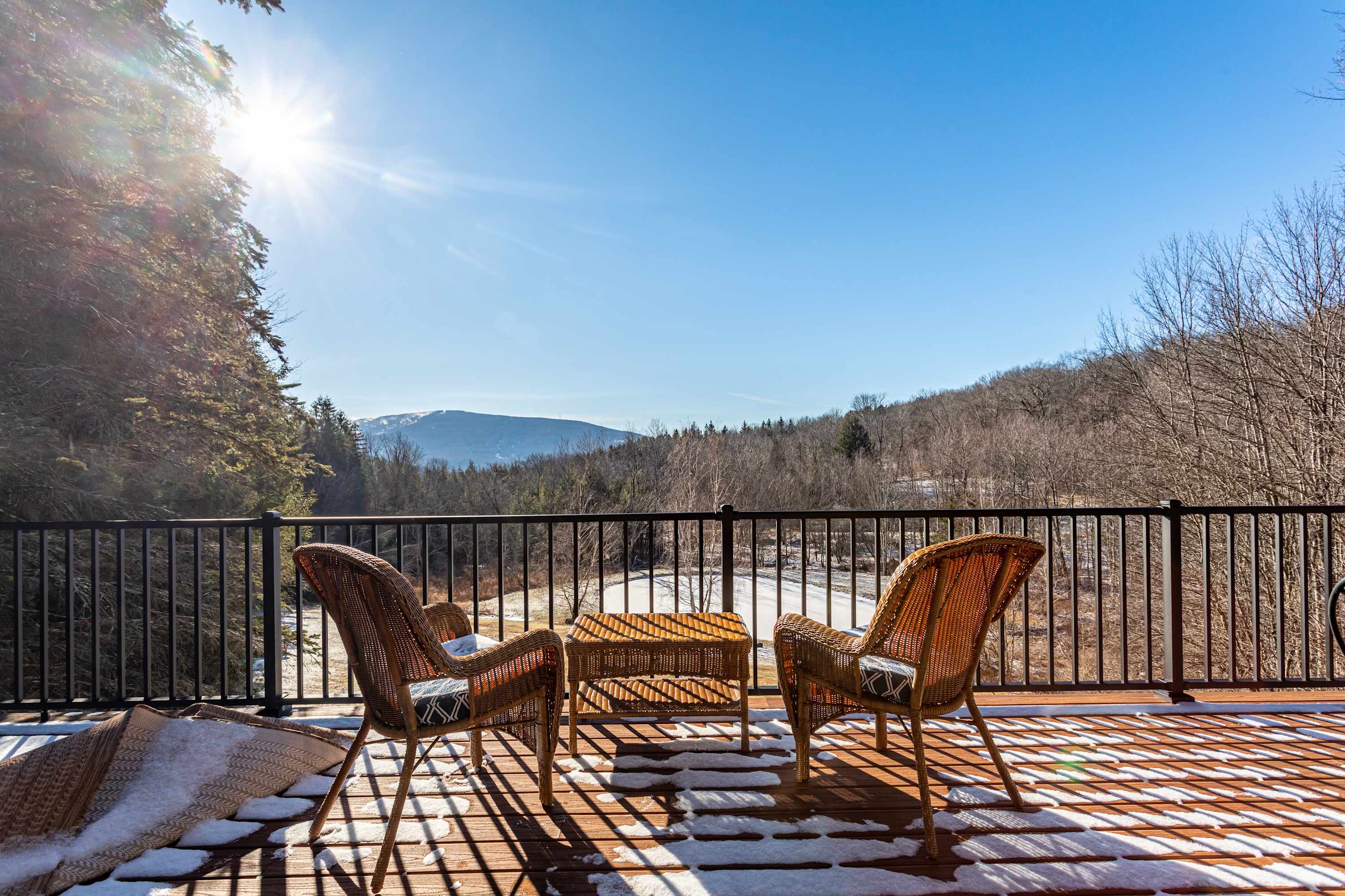 A sunny outdoor deck with wicker chairs and a small table, offering expansive views of a snow-dusted valley and distant mountains under a clear blue sky.